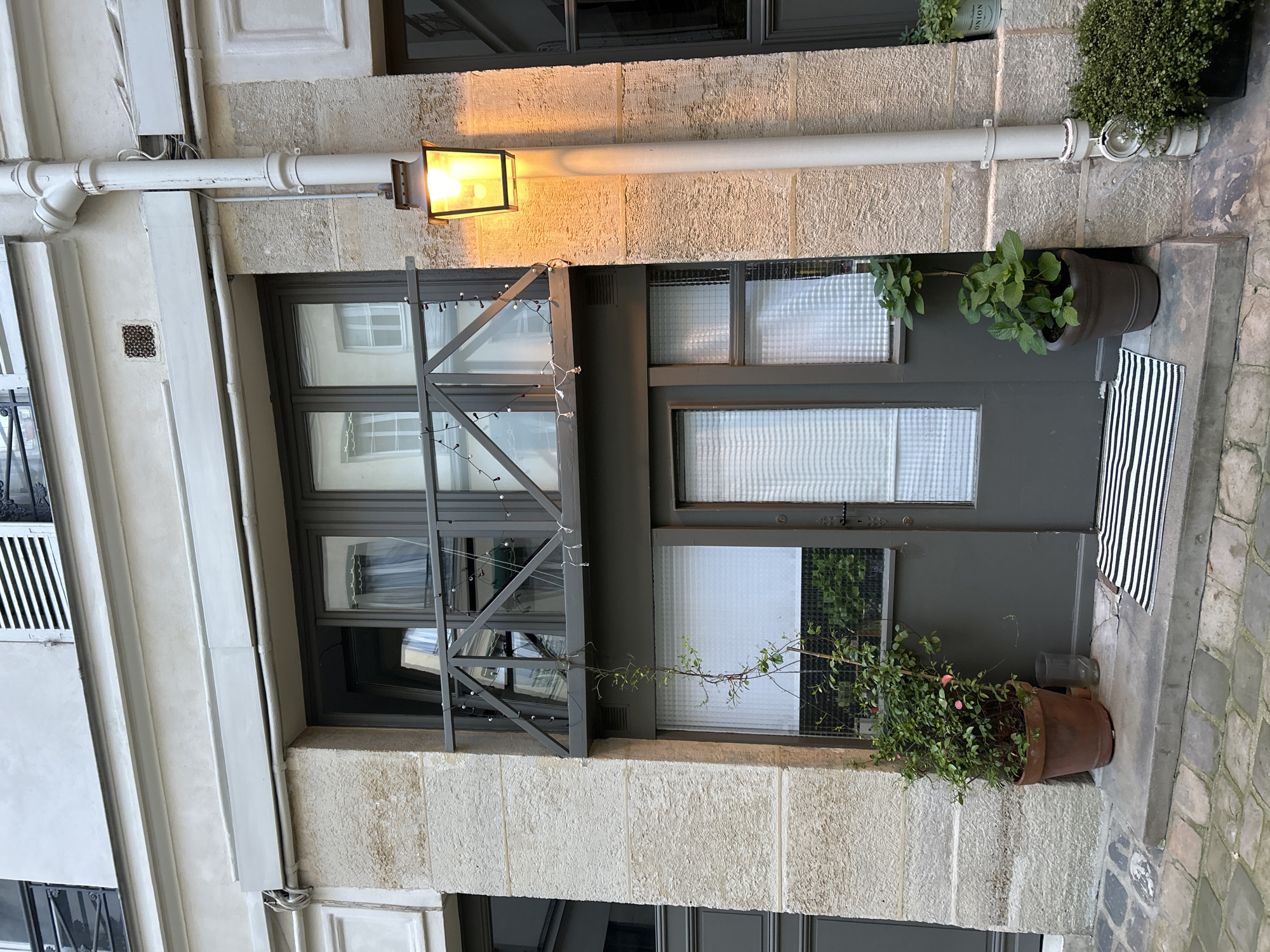 This image shows a gray-colored window and door on a stone building, likely in Europe. A white pipe with a light fixture runs horizontally above the window and door. The window is divided into panes with a gray frame, and the door has a similar design. A decorative metal structure with small lights adorns the window. Potted plants are placed on either side of the door, and a striped mat is visible near the entrance. The street is paved with cobblestones.