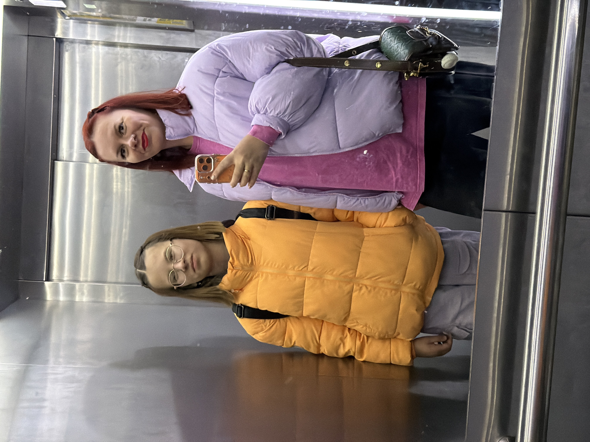 Two women are reflected in what appears to be a mirrored wall or elevator in an indoor location. The woman at the top of the frame has red hair and is wearing a light purple puffer jacket over a pink top. She holds an orange-cased smartphone, seemingly taking a photo or video. Her handbag, a dark green color with a brown strap, is visible over her shoulder. The woman at the bottom of the frame wears round glasses and an orange puffer jacket with black straps. She appears to be looking at the viewer or reflection. Both women are wearing jackets, suggesting a cool temperature. The background is a plain, reflective surface.