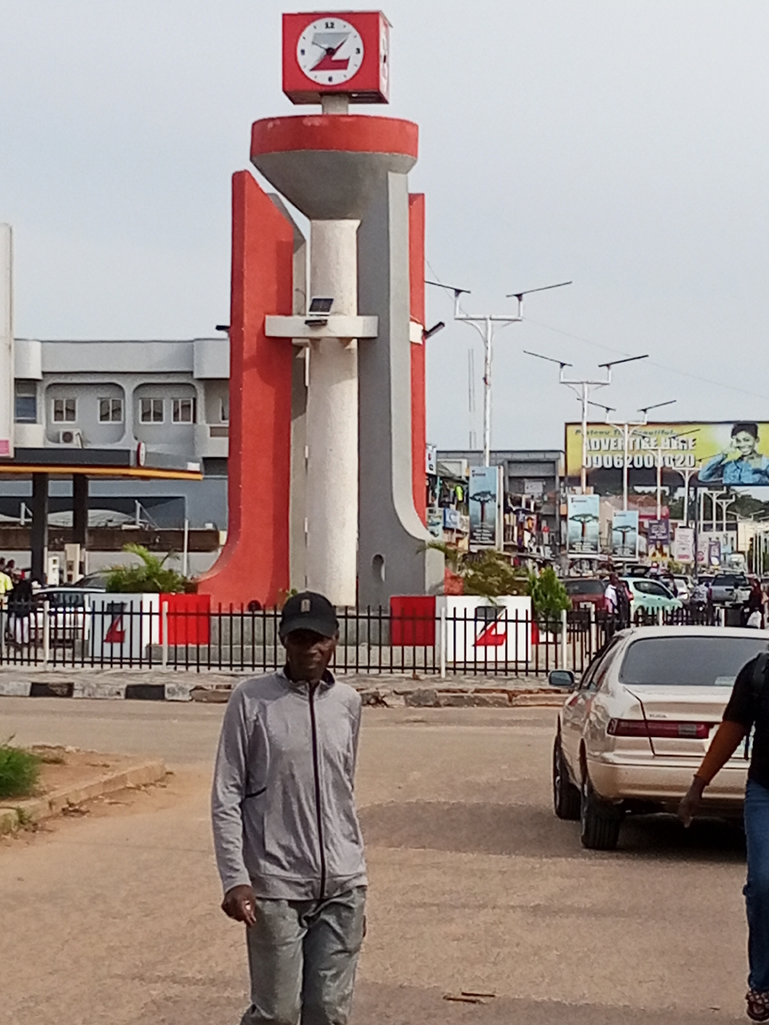A man walks towards the viewer on a paved road in front of a large, red and grey clock tower. The clock tower is a prominent landmark, with a red-capped top featuring a white clock face with black Roman numerals and hands. The tower itself has a grey cylindrical body encased in red, curved panels. In the background, a street scene unfolds with buildings, other streetlights, traffic, and advertisements. A beige sedan is parked to the right, and the man is walking in the foreground, wearing a grey zipped jacket and grey pants. The sky is overcast.