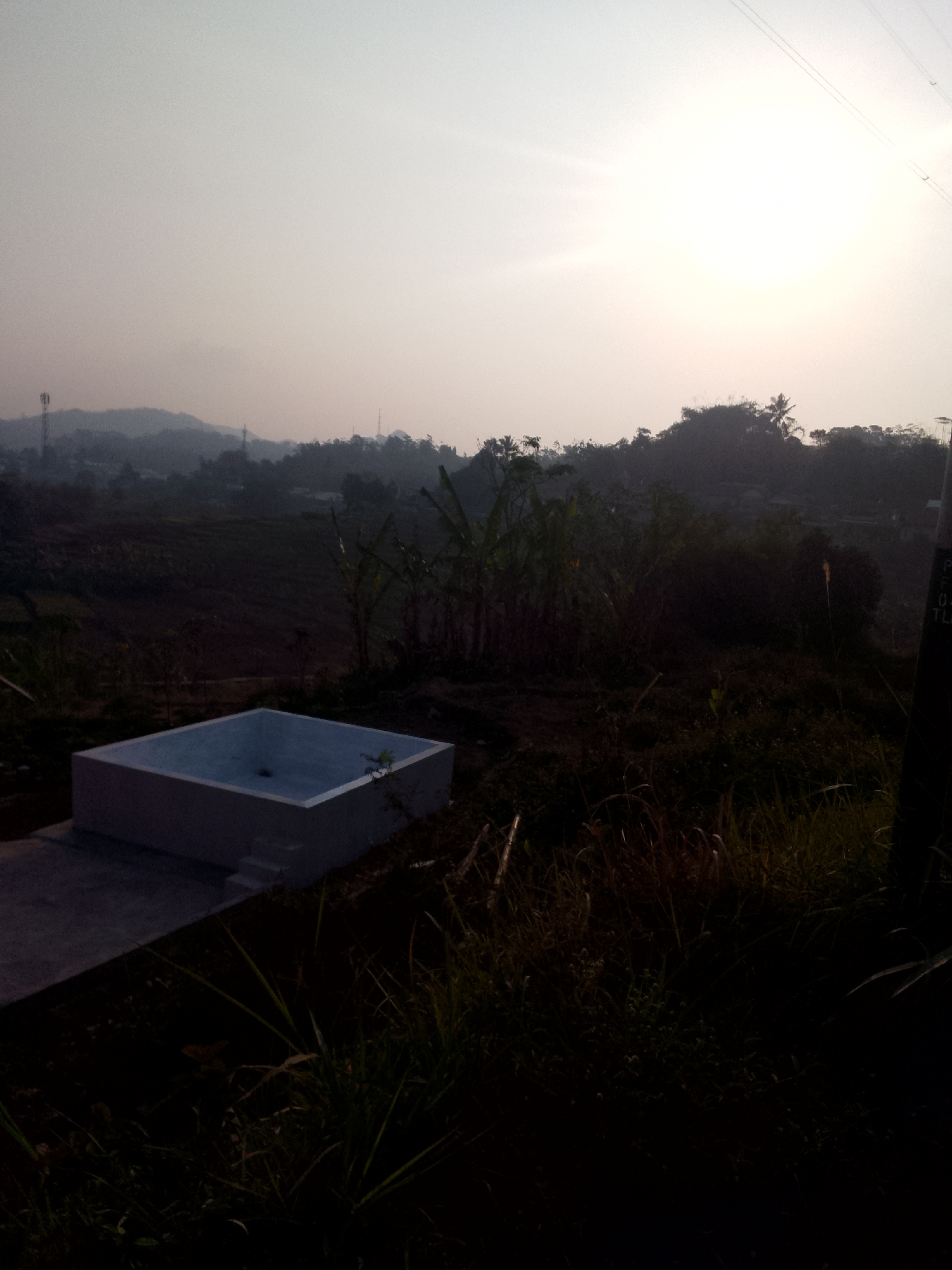 The image captures a serene, early morning scene in Padalarang, Indonesia, with a view of rolling hills and lush vegetation under a hazy sky. In the foreground, a modern, light-colored, rectangular swimming pool with a few steps leading into it is situated on a concrete platform. The pool appears empty. Surrounding the pool is dense, dark foliage, including tall grasses and some banana trees, suggesting a semi-rural or undeveloped area. In the mid-ground, the landscape transitions to terraced fields, indicating agricultural activity. Distant hills form the backdrop, with a faint haze obscuring their details. A telecommunication tower is visible on the left side of the frame, silhouetted against the sky. The overall atmosphere is quiet and tranquil, illuminated by the soft light of an approaching sunrise or sunset, casting a diffused glow over the scene. The image does not contain any people or signs of recent activity, emphasizing a sense of stillness.