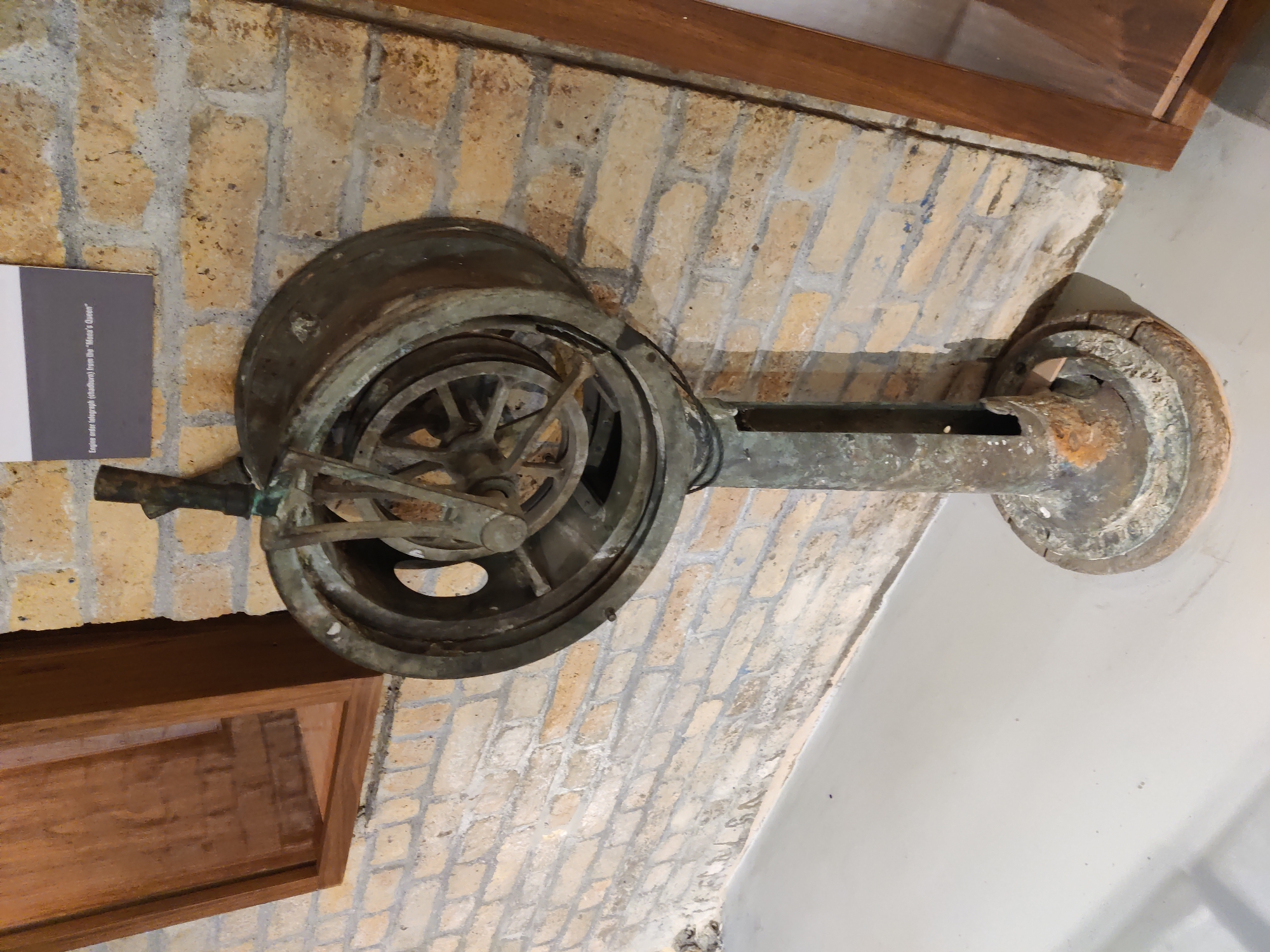 An old, rustic engine order telegraph, likely made of brass or bronze, stands upright on a light gray floor against a light brick wall. The telegraph has a circular mechanism at the top with spokes and what appears to be a handle or lever extending from it. Below the circular mechanism is a long, cylindrical stand that tapers to a wider, circular base. The entire object shows signs of age and wear, with rust and discoloration visible. To the left of the telegraph is a wooden cabinet or display case, also appearing aged. A small sign is mounted on the brick wall above and to the right of the telegraph, with text that reads "Engine order telegraph (chadburn) from the 'Mona's Queen'". This object is a historical artifact, likely on display in a museum or exhibition. The environment suggests an indoor setting, possibly a historical exhibition hall, given the display of artifacts and informative signage. The location is identified as Dunkirk, France, which hints at the potential historical context of the artifact, possibly related to maritime history given the nature of an engine order telegraph.