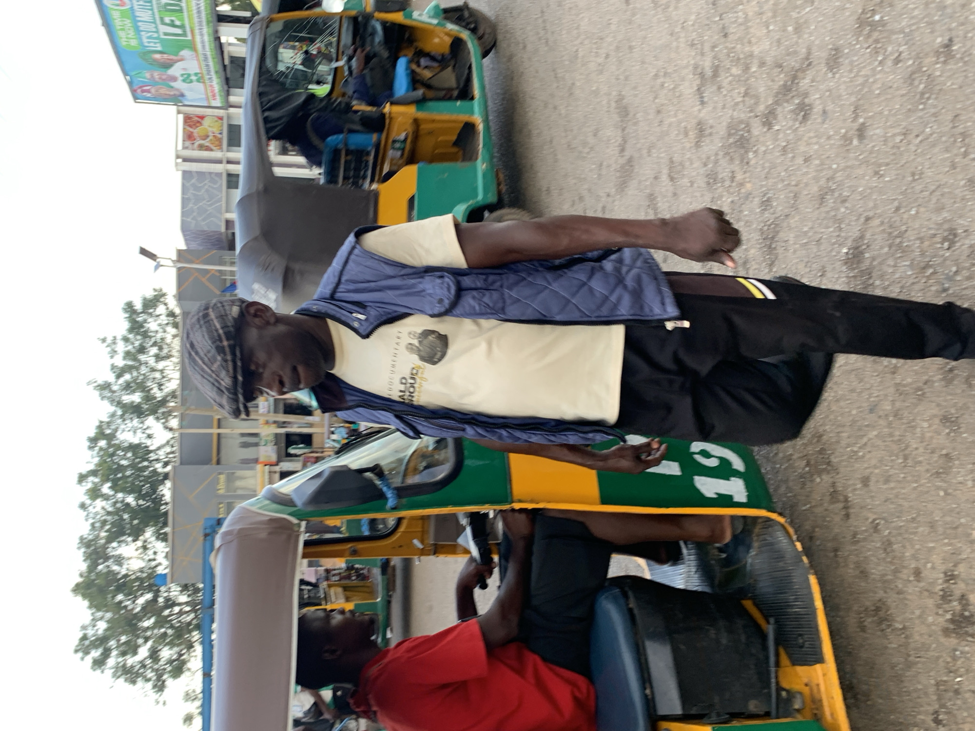 The image captures a street scene in Jos, Nigeria, with a focus on two men and a trishaw, also known as an 'Okada'. In the foreground, a man wearing a blue quilted vest over a light-colored t-shirt and black pants walks towards the right of the frame. He has a flat cap on his head. To his left, the driver of a green and yellow trishaw, dressed in a red t-shirt and black shorts, is seated behind the handlebars, appearing to be in motion or waiting. The trishaw has the number "19" painted on its side. In the background, other vehicles, including another trishaw, are visible, along with buildings and trees. A large billboard displaying political imagery and text, including "THE TIME IS NOW. LET'S DO MUTUAL", is prominent in the upper right portion of the image. The ground is paved with asphalt.