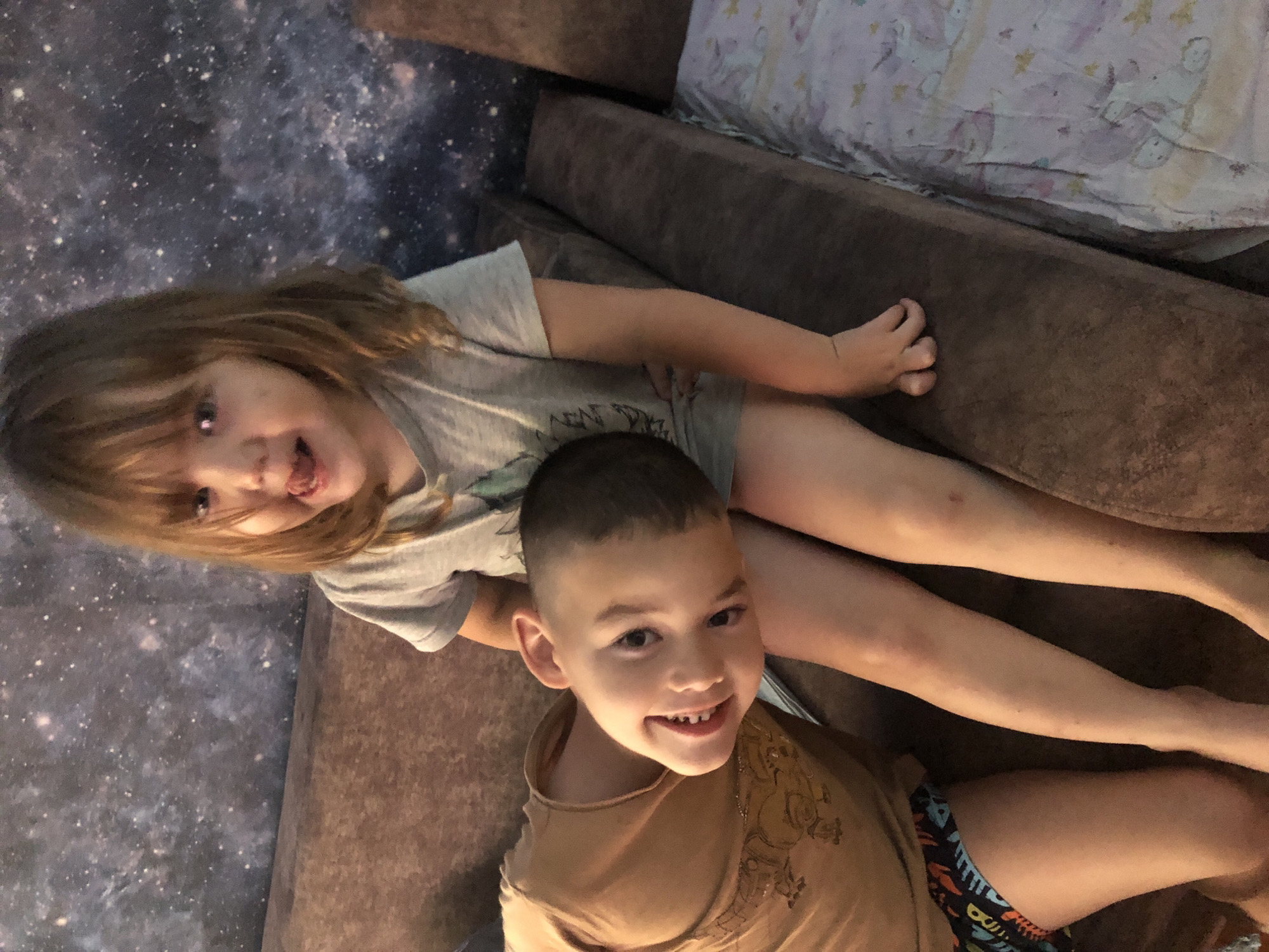 Two young children are sitting on a brown couch in front of a dark wall with a starry pattern. The child on the right, a girl with long brown hair, is wearing a grey t-shirt and is sticking her tongue out. The child on the left, a boy with short brown hair, is wearing a tan t-shirt with a graphic and patterned shorts. He is leaning his head on the girl's leg and smiling. The couch appears to be in a domestic setting. The location in Tashtyp, Russia is not determinable from the image.