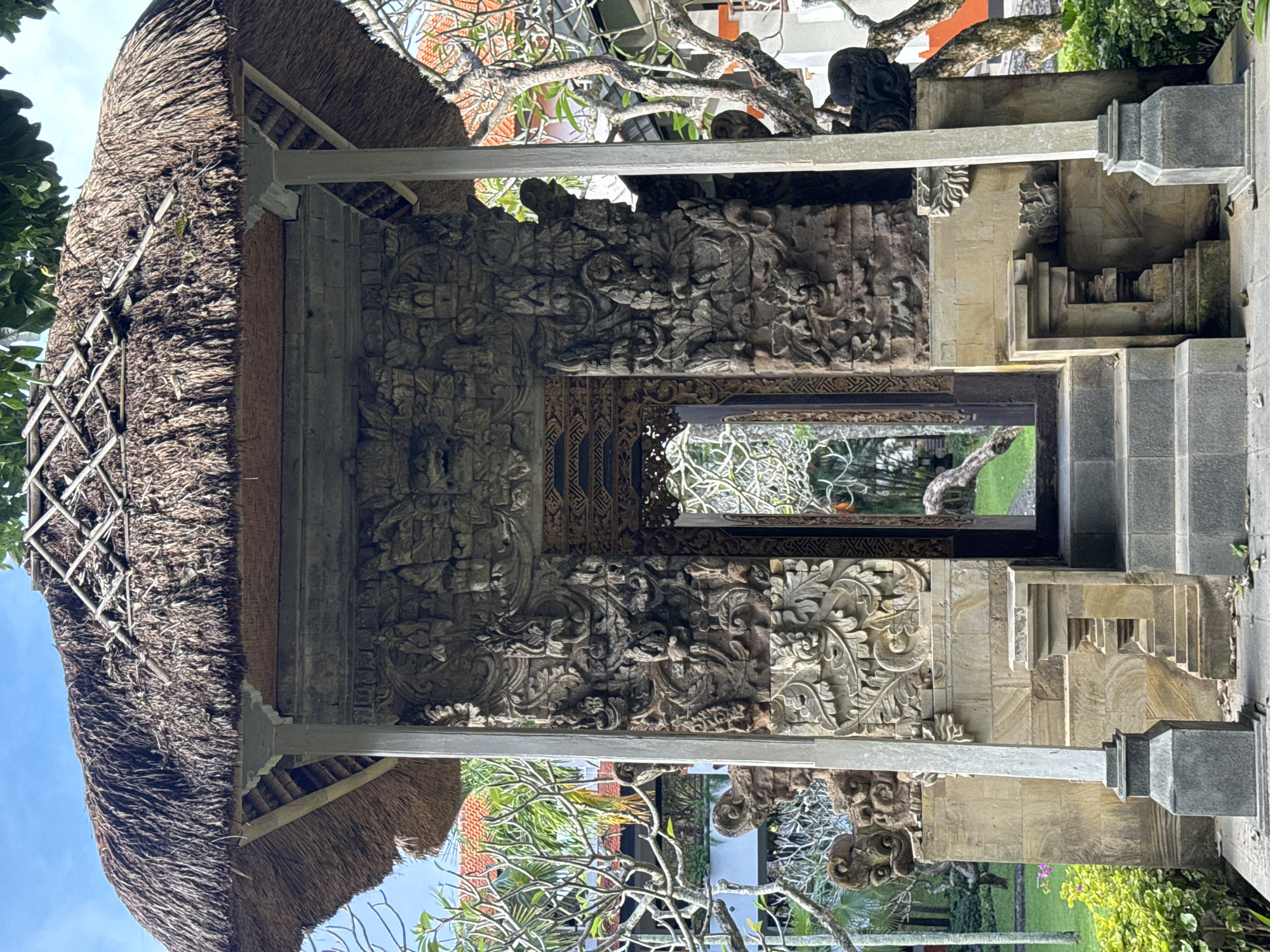 The image depicts a Balinese gate structure, likely located in Kuta Selatan, Indonesia, given the style of architecture and carvings. The gate features a thatched roof with intricate carvings on the stone walls, depicting floral and mythical figures. A passage is visible through the gate, revealing greenery and possibly reflecting the surrounding landscape. Steps lead up to and through the gate, suggesting a transition point between spaces. The structure is surrounded by trees and other vegetation. No people are visible in the immediate vicinity.