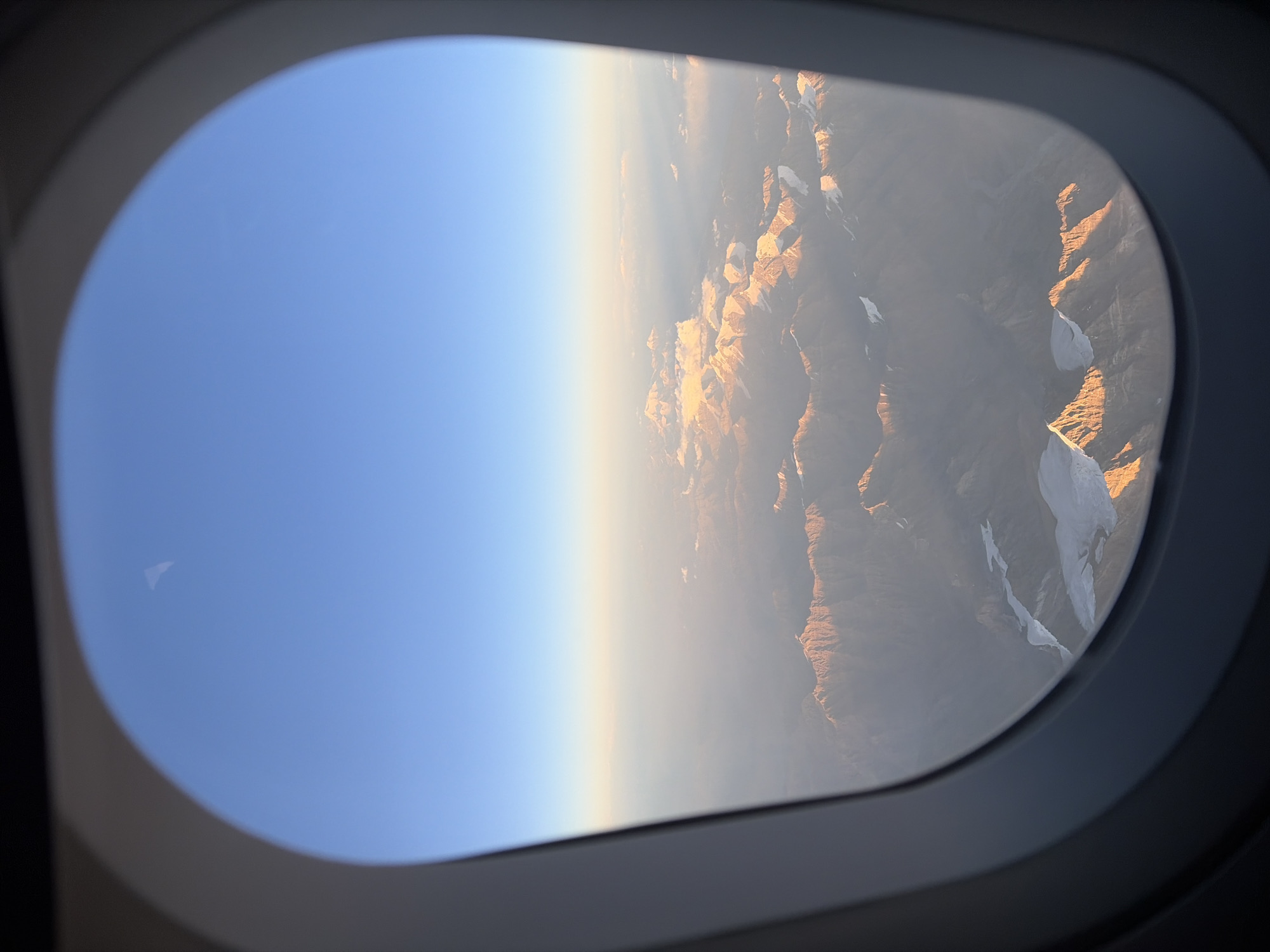 The image shows a view of snow-capped mountains from an airplane window. The sky is a clear blue, with a gradient of lighter blue and orange near the horizon, suggesting either sunrise or sunset. The mountains are rugged and appear to be covered in snow and ice, with some areas showing rocky terrain. Sunlight is illuminating the peaks, creating a warm glow and casting shadows. In the foreground, the dark frame of the airplane window is visible. The overall scene depicts a vast, mountainous landscape from an aerial perspective. The location is identified as Unknown, Tajikistan.