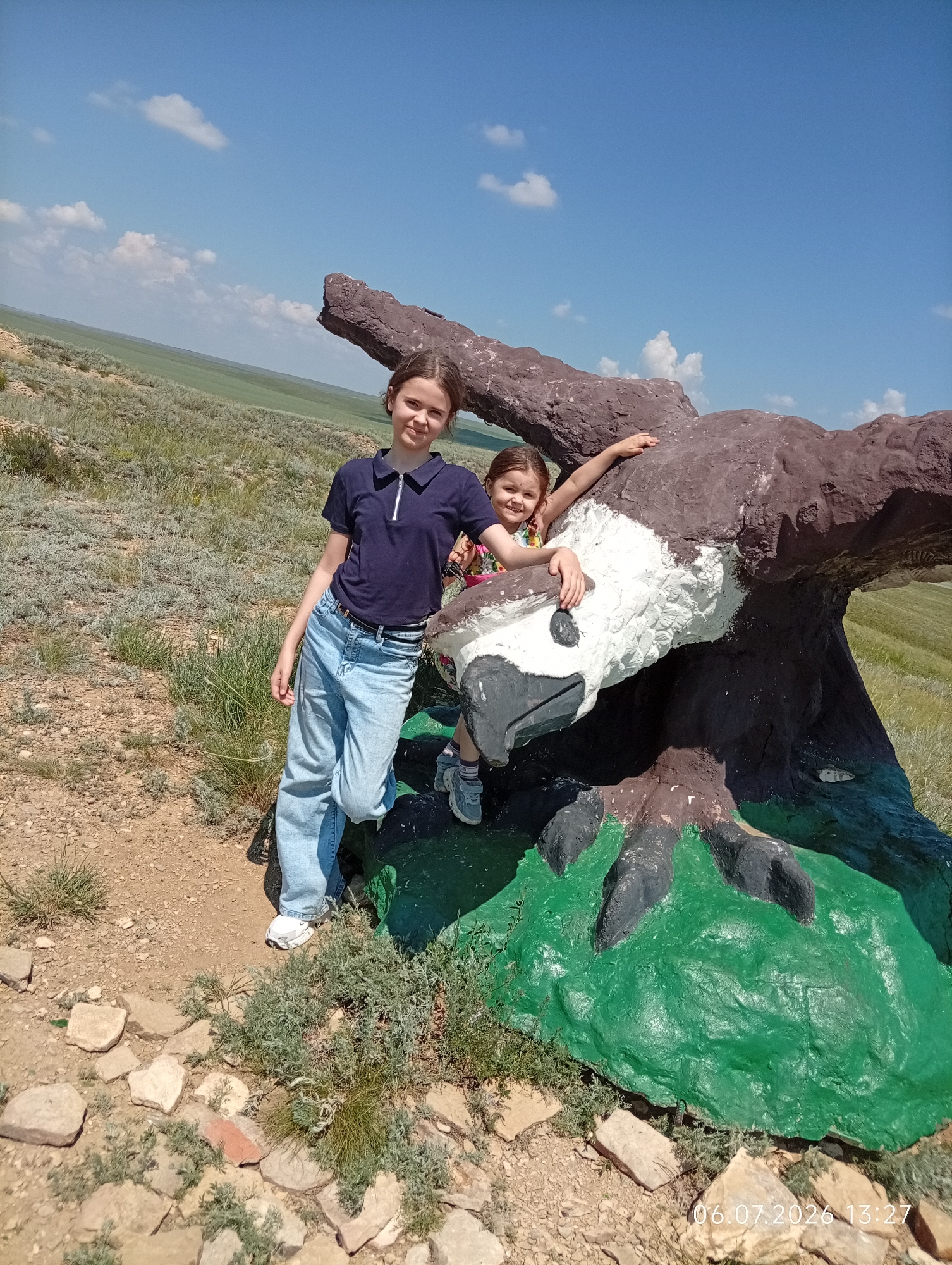 Two young girls are posing for a photograph outdoors, in a hilly, grassy landscape under a bright blue sky with scattered white clouds. The girl on the left, appearing older, stands facing the camera, leaning slightly on a large, painted sculpture of a bird of prey, possibly an eagle. She is wearing a dark blue polo shirt and light blue jeans. The younger girl is peeking out from behind the sculpture, smiling at the camera. She is wearing a floral-patterned shirt. The sculpture itself is roughly painted with brown, white, and black, with a bright green base. The ground is a mix of dirt, rocks, and sparse vegetation. In the background, the landscape stretches out with rolling hills covered in dry grass. A timestamp in the lower right corner reads "06.07.2026 13:27". The location is identified as Aleksandrovka, Kazakhstan.