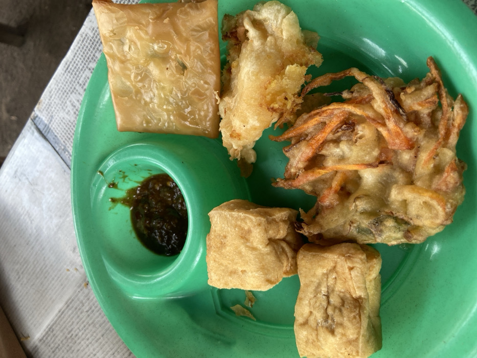 A close-up, elevated view captures a bright green, plastic plate containing various fried Indonesian snacks and a condiment. The plate features an integrated circular well on the upper left, which holds a dark green-brown, viscous sauce or sambal with minor splatter marks on its rim. To the immediate left of the well are two irregular, light brown, rectangular pieces of fried tofu (tahu goreng). Below them is one irregular-shaped, golden-brown vegetable fritter (bakwan or bala-bala) displaying visible strands of orange (likely carrot) and other fragmented green vegetables. To the right of the sauce well is a square, golden-brown, flaky fried pastry, possibly a type of martabak or spring roll. Below this, to the right of the tofu, lies an irregular, light golden-brown fried item, possibly tempeh mendoan or fried mushroom. The plate rests on a surface covered by a patterned, greyish-white material, consistent with newspaper, visible as a blurred background element. No individuals are present. The scene is located in the city of Pandanarum, Indonesia.