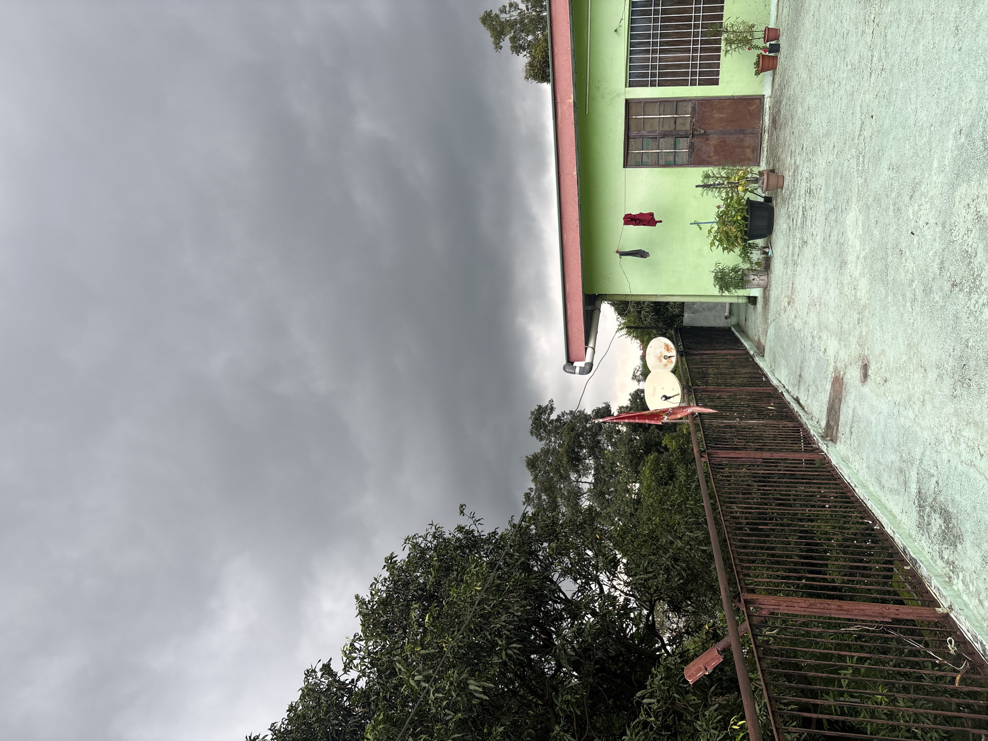 The image depicts a rooftop overlooking a verdant landscape with a light green building in the foreground. The sky is overcast with dark, heavy clouds, suggesting impending rain. The building, painted a vibrant light green with a red trim, features two brown wooden doors and a window with white bars. Several potted plants are arranged on the rooftop in front of the building. A rusty metal railing runs along the edge of the rooftop, separating it from the dense foliage below. Two satellite dishes and a red flag are visible near the railing. Two articles of clothing, a dark item and a red item, are hanging on a clothesline attached to the building. The scene suggests a residential setting in Baldwara, India, under a brooding sky.