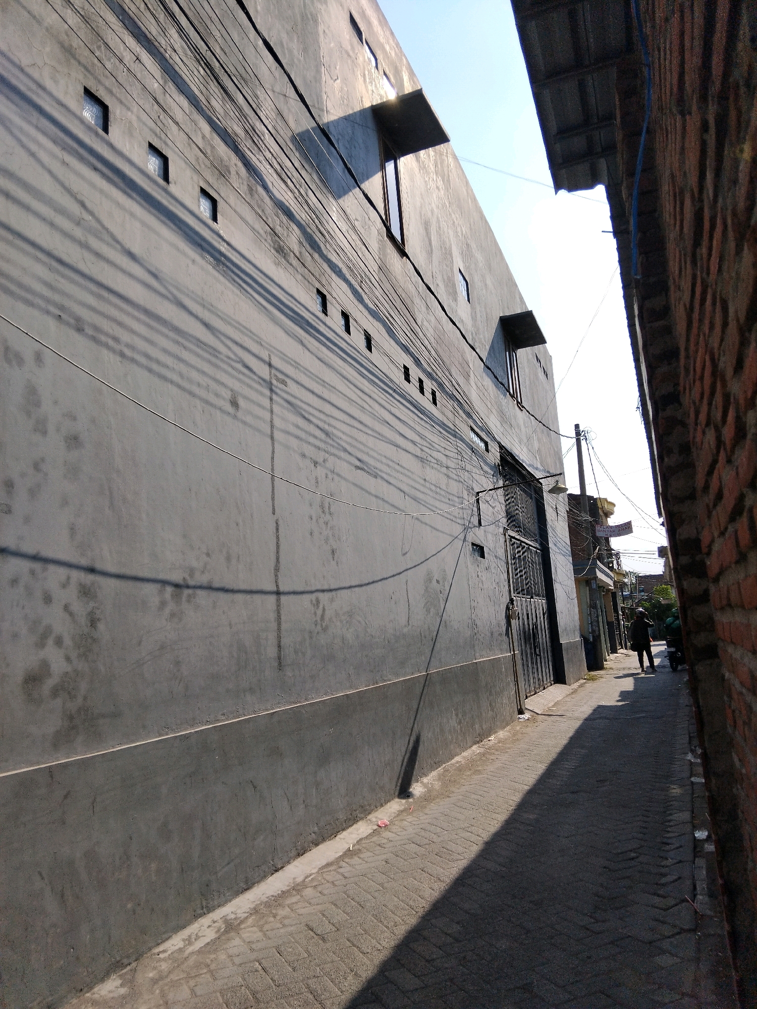 The image depicts a narrow alleyway in the city of Kepuh Kiriman, Indonesia. The alley is flanked on the left by a tall, grey, concrete building with multiple small, square windows. Numerous electrical wires crisscross the face of this building, casting long shadows on its surface. The building has a textured, weathered appearance with some water stains. On the right side of the alley is a brick building, partially visible. The alley floor is paved with interlocking grey bricks.
In the distance, a person is walking away from the camera, silhouetted against the bright sky. A motorcycle is parked near this person. The alley is a sunlit area, with strong shadows indicating a bright day. There is debris, including small red and white objects, scattered along the edge of the alley pavement. The overall impression is of a utilitarian and somewhat aged urban environment.