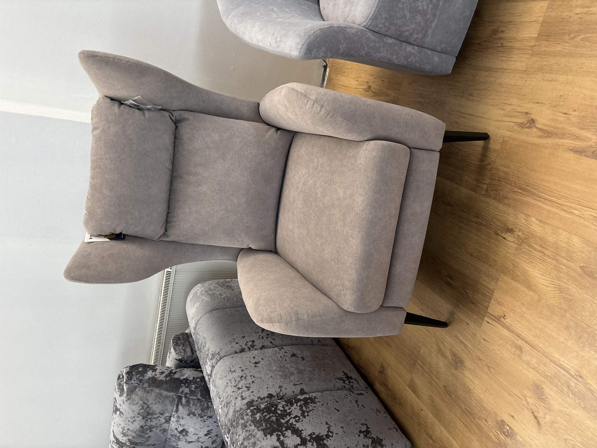 This media file depicts a top-down, rotated view of several pieces of upholstered furniture. The primary subject is a large, light grey armchair with plush cushioning. The armchair features a high backrest, wide armrests, and a separate headrest or lumbar support pillow. A small, clear plastic tag is attached to the upper corner of the armchair's backrest. To the lower right of the armchair, a dark, slender furniture leg is visible, resting on a light brown, wood-grain patterned floor.
In the background, a portion of another grey armchair, with a textured, possibly velvet-like finish, is visible. Further into the background, another piece of furniture, also grey with a similar textured finish, is present, showing a curved armrest. The setting appears to be indoors, possibly a showroom or a living space, given the presence of furniture and the polished floor. The lighting is bright, suggesting daytime or well-lit indoor conditions. There is no discernible weather information. No people are visible in the frame. The image is a still photograph.