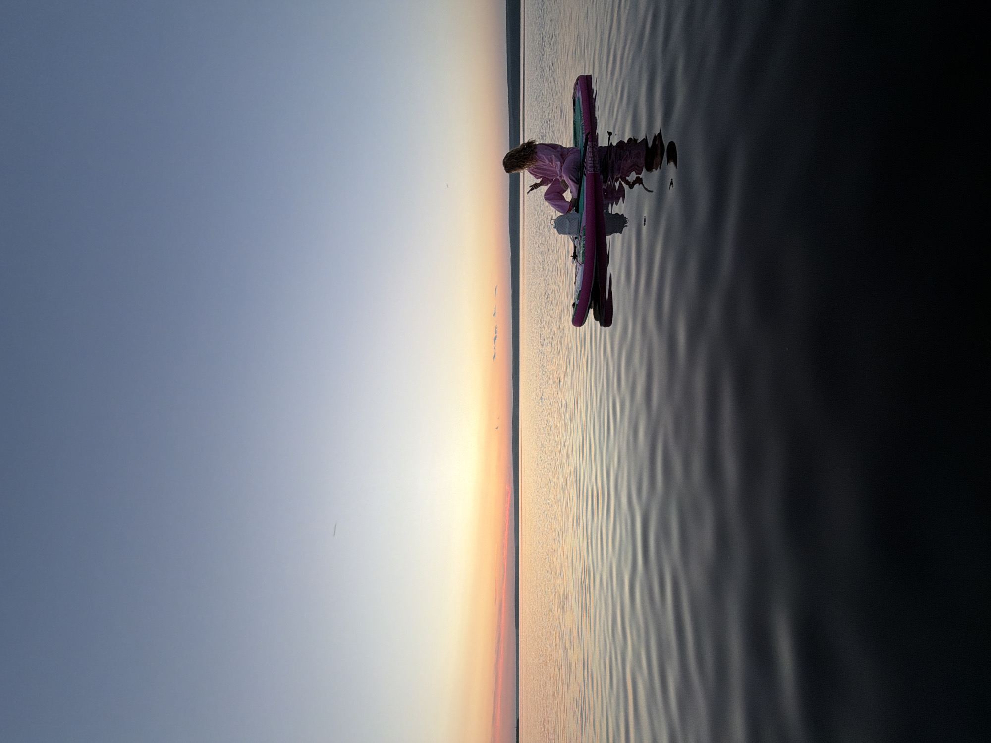 A woman in pink loungewear sits on a pink stand-up paddleboard in the calm waters of the Kama River during sunset. The sky displays a gradient of colors from pale blue at the top to orange and pink near the horizon. A thin, dark landmass is visible in the distance, separating the sky from the water. The water's surface has gentle ripples, reflecting the sky and the paddleboard. A small, light-colored backpack rests on the paddleboard beside the woman, who is holding a smartphone. The scene is serene and bathed in the soft light of dusk.