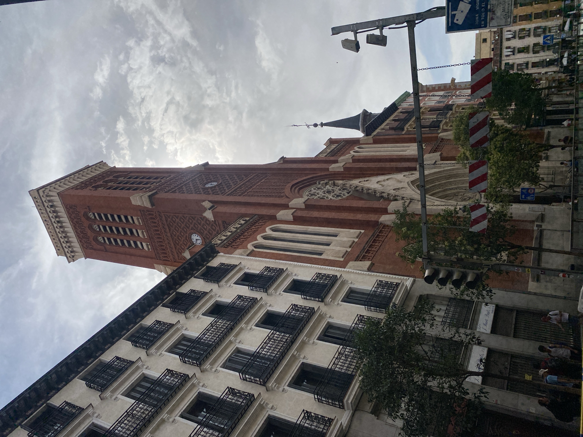 The media file displays a view of urban architecture under a cloudy sky. The primary subjects are two buildings: a tall, red brick church with a clock tower and ornate windows, and a lighter-colored apartment building with multiple floors, each featuring numerous windows with black wrought-iron balconies. Trees are visible between and beside the buildings, adding a touch of nature to the urban scene. Several red and white striped warning signs hang from a metal pole in the foreground, suggesting ongoing work or a temporary hazard. A traffic light with four bulbs is also attached to the pole.
In the background, more apartment buildings can be seen, as well as street signs. A few people are faintly visible on the street, suggesting a typical day in a city. The weather appears overcast with some breaks in the clouds, indicating a diffused daylight, possibly late morning or early afternoon.
The overall scene is one of everyday urban life in Madrid, Spain, characterized by historic architecture and modern residential buildings. There are no explicit activities or events depicted, but the presence of people and traffic signals implies a functioning urban environment. Visible text includes "ZONA VIGILANCIA" and "AYUNTAMIENTO DE MADRID" on a blue sign, indicating a surveillance zone in the city of Madrid. Another sign appears to list "J. MACK PRATS, S.L."