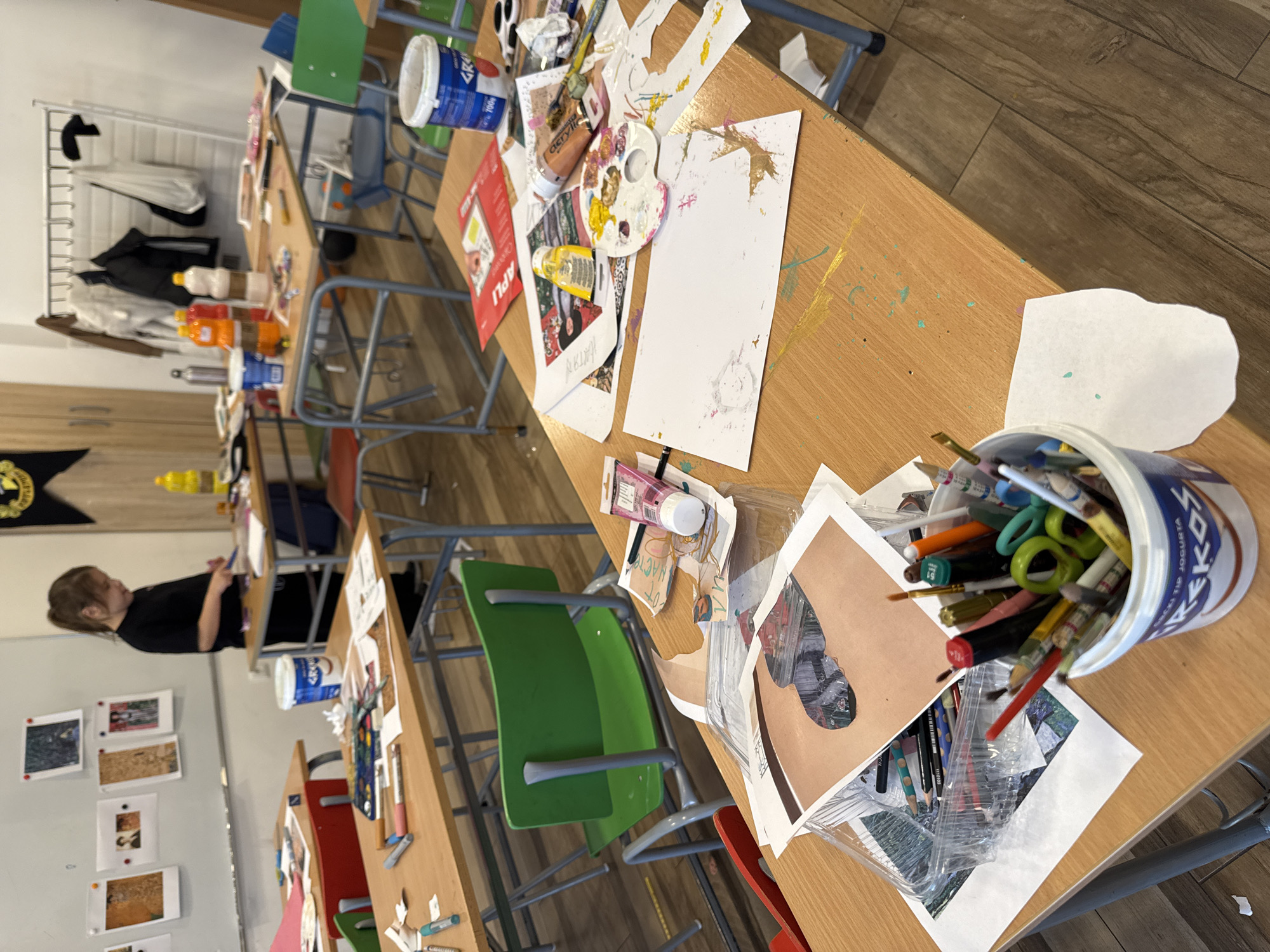 The image shows a classroom setting, possibly in Budva, Montenegro (as indicated by the writing on a bucket), with several rows of desks and chairs. The desks are covered with art supplies, including brushes, pencils, paints, paper, and various containers. A person with short brown hair is visible in the background. The room appears to be an art studio or classroom, based on the abundance of art-related materials and the presence of artwork displayed on the wall.