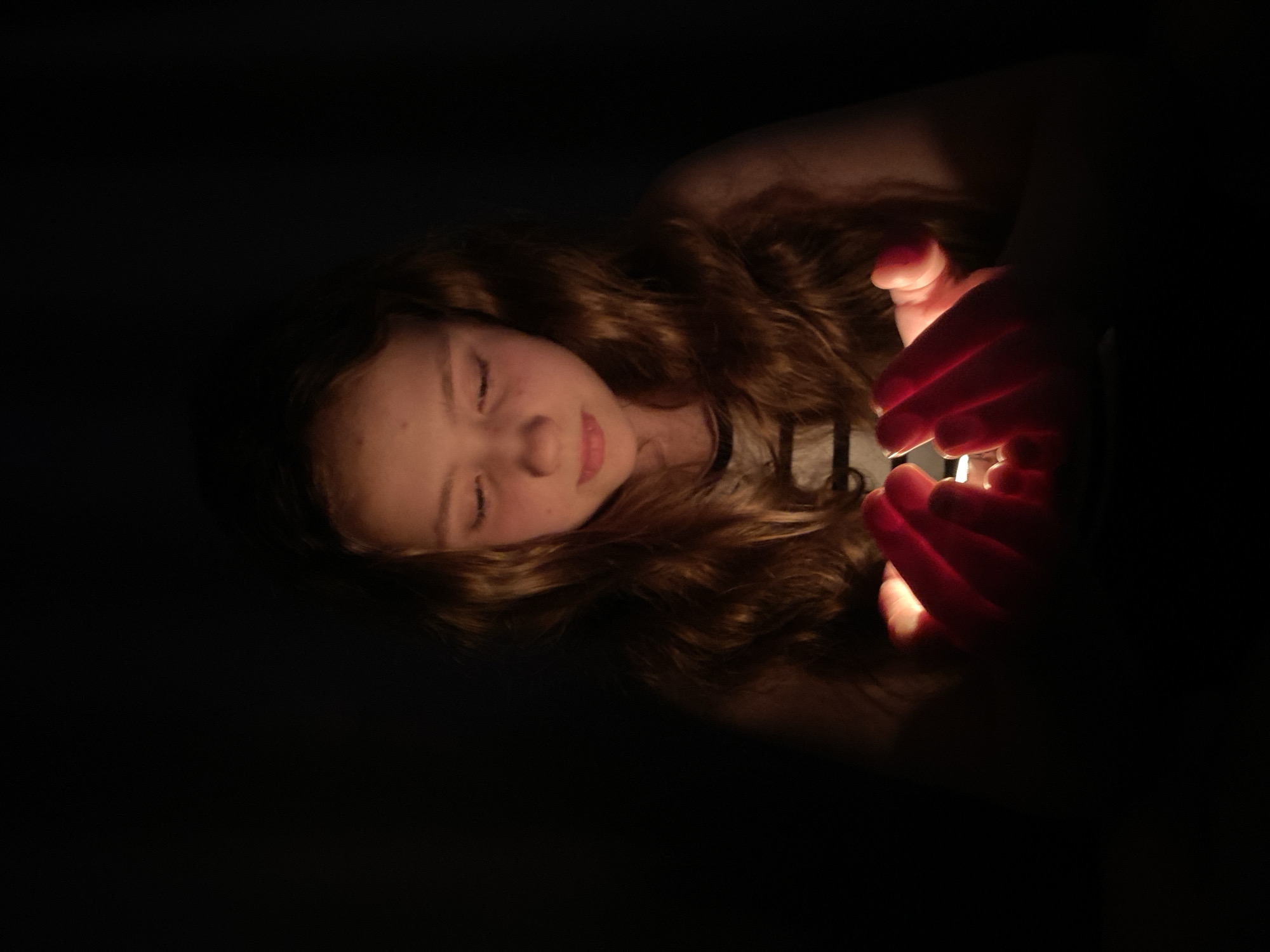A dimly lit image shows a young girl lying down with her eyes closed. She has long, wavy brown hair and is wearing a light-colored top with a dark neckline. Her hands are cupped together, glowing with light, suggesting she is holding a small light source such as a candle or a phone. The background is dark, and the location appears to be indoors.