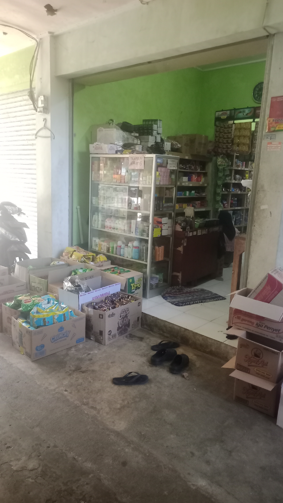 The image shows the exterior of a small store in Cogreg, Indonesia, with several cardboard boxes stacked on the ground in front of it. The store appears to be a convenience store or minimarket, with shelves stocked with various products visible through the open doorway. The boxes on the ground contain packaged goods, possibly snacks or other retail items. Some of the boxes have visible branding, including "Crunchy" and "Taro" snacks. Several pairs of black flip-flops are also scattered on the ground. The building has a light-colored facade and a green-painted interior wall visible. A motorcycle is parked to the left of the store entrance. The overall scene suggests a typical street-level retail environment in a residential or commercial area.