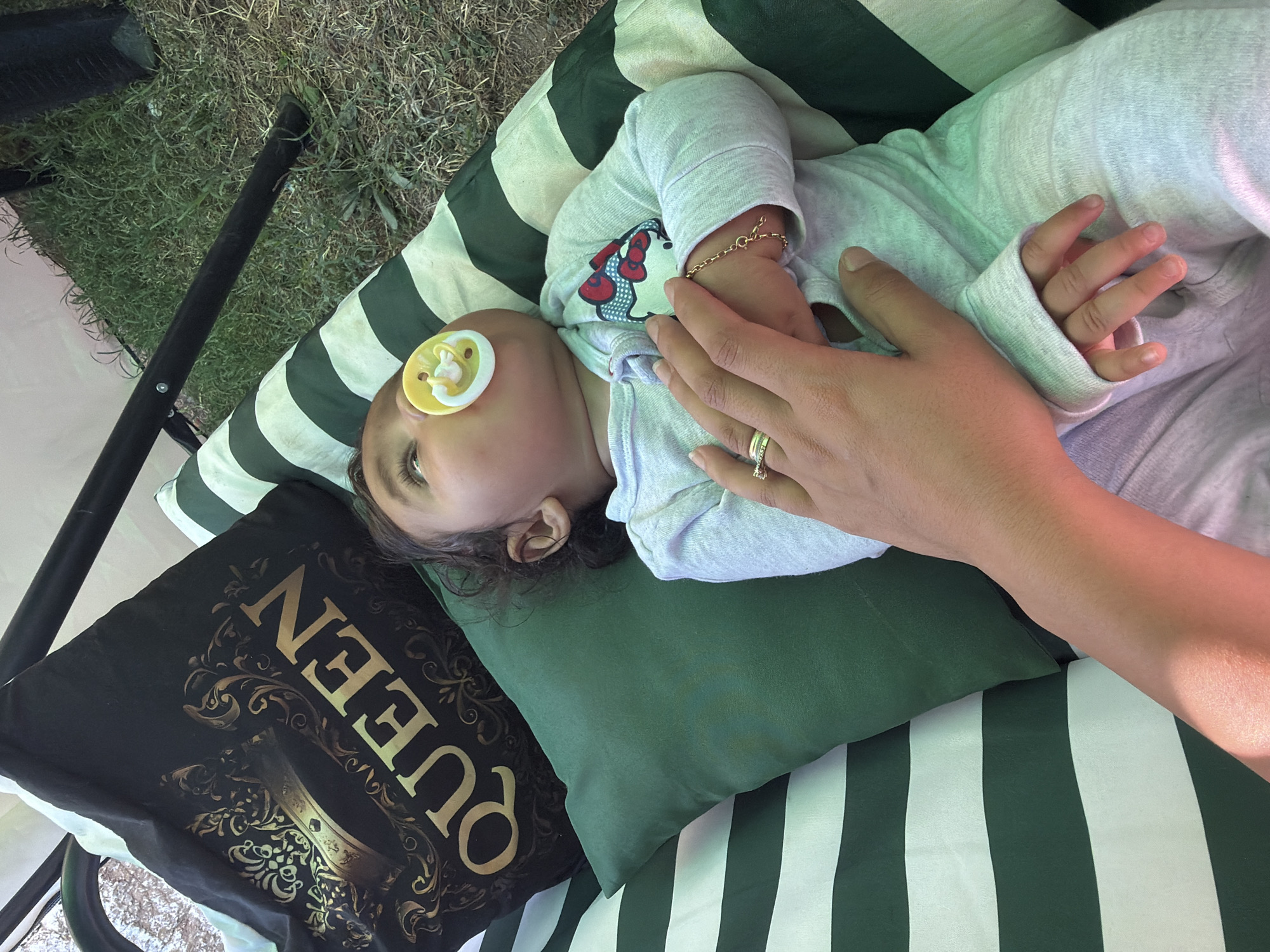 An infant is lying on a striped green and white cushion, propped up against a black pillow with the word "QUEEN" in gold lettering. The infant has a yellow pacifier in its mouth and is wearing a light gray onesie with a Hello Kitty graphic on the chest. A person's hand, wearing a ring, rests on the infant's chest, and a delicate gold bracelet is visible on the infant's wrist. The scene appears to be outdoors, with a patch of green grass and a dark metal swing frame visible in the background, suggesting it is in Razhevo Konare, Bulgaria, in a yard or garden setting.