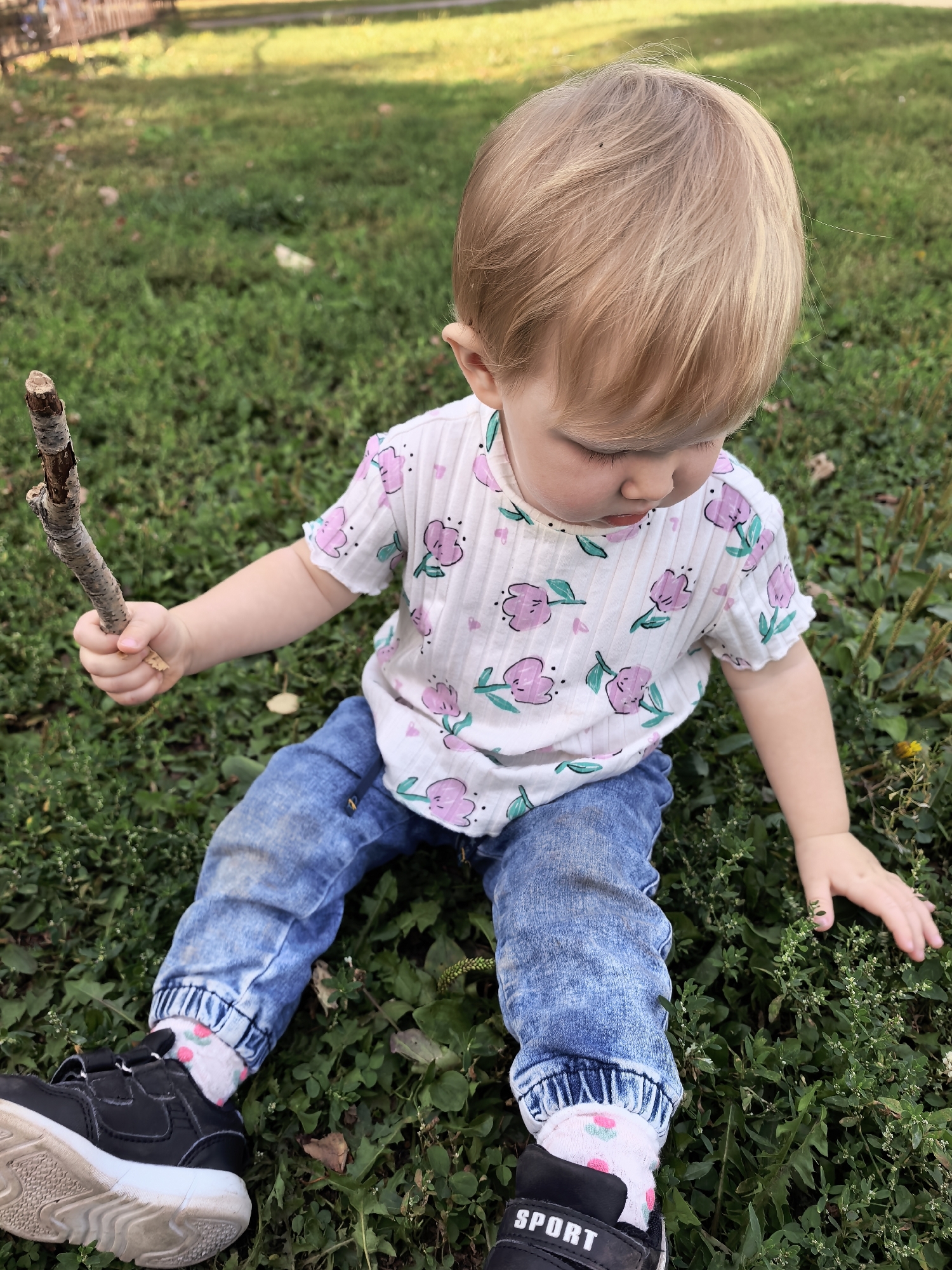 A young child with blond hair sits on a grassy area, holding a stick. The child is wearing a white t-shirt with pink floral patterns, blue jeans, and black sneakers with white soles and pink socks with a floral pattern. The child's left hand holds the stick, and the right hand is resting on the grass. The background shows more grass and a blurred fence or structure, suggesting an outdoor setting. The image appears to be taken during the day, with natural light. There is no identifiable information within the image to suggest the location is Sterlitamak, Russia.