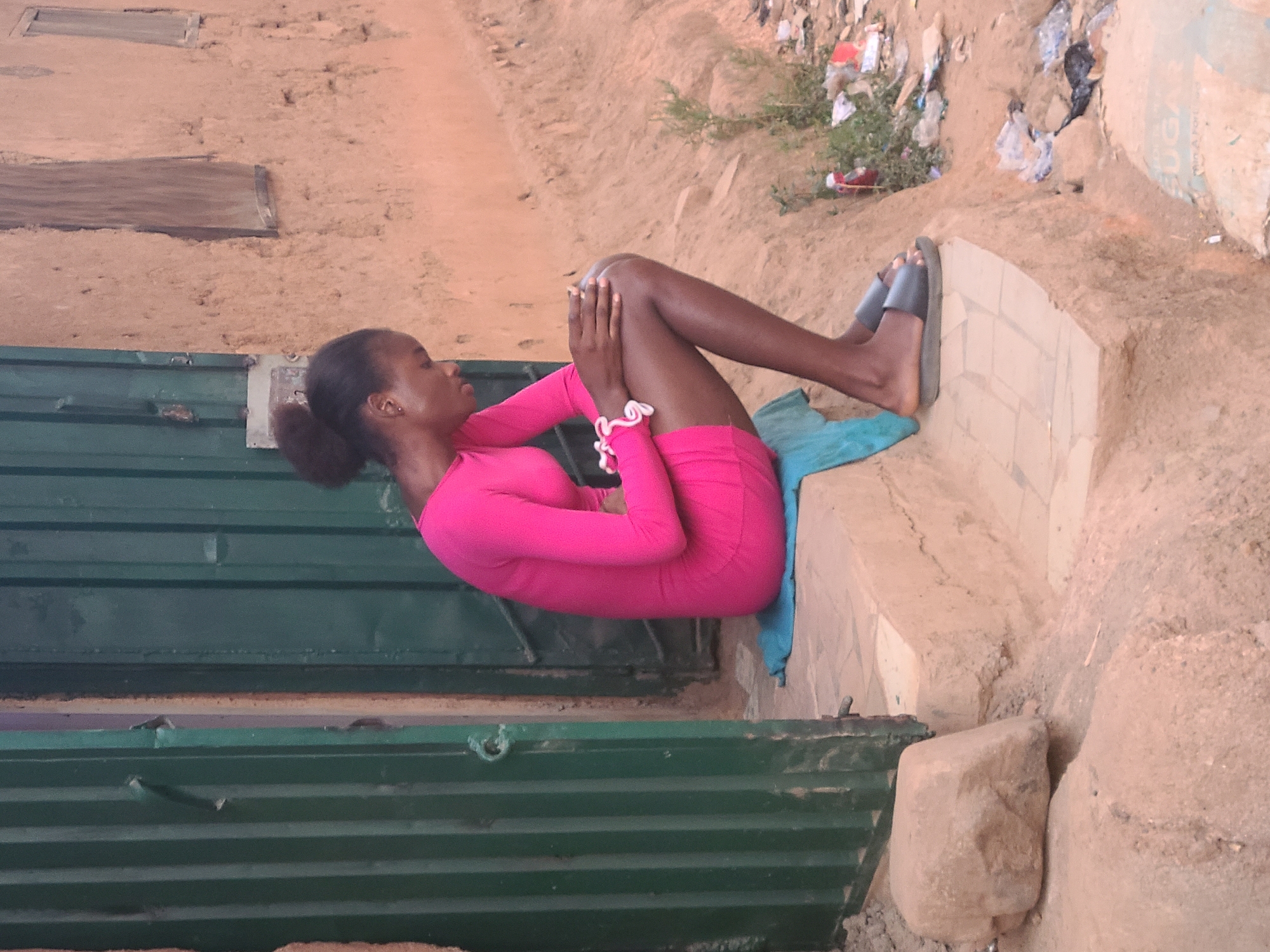 A young woman in a bright pink, long-sleeved dress is sitting on a concrete step, propped up by a folded blue towel. She is wearing dark-colored sandals. She is hugging her knees to her chest with her hands interlocked, and she is facing left. Her hair is pulled back in a low bun. She is in front of a corrugated green metal structure with horizontal ridges. The setting appears to be outdoors, with a sandy, uneven ground and litter visible in the background. The photo was taken in Jos, Nigeria.
