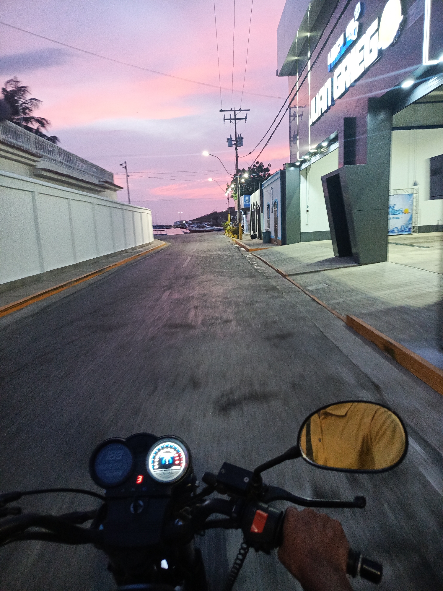 A point-of-view shot from a person riding a motorcycle down a paved street in Juan Griego, Venezuela. The sky is a mix of pink and purple hues, indicating either sunrise or sunset. On the right side of the street is a modern, multi-story building with a prominent sign reading "LUAN GREG," lit from underneath. Along the left side of the street is a high, white wall. Streetlights are visible in the distance. The motorcycle's dashboard displays a speedometer reading 188 and a tachometer at 3. The rider's hand is visible on the throttle. The mirror reflects the rider wearing a yellow shirt. In the distance, the street leads to what appears to be a coastal area with boats. Power lines stretch across the sky above the street.