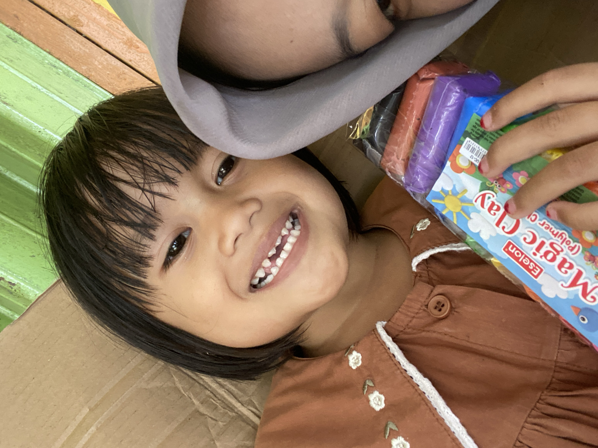 An outdoor close-up selfie shows a young girl with dark hair and a brown dress smiling at the camera, holding a package of "Magic Clay" with her fingers. She is positioned on the left side of the frame. To her right, partially obscured by a grey hijab, is another person's face. The background is a textured brown cardboard surface and a green wooden wall. The scene is set in Macege, Indonesia.