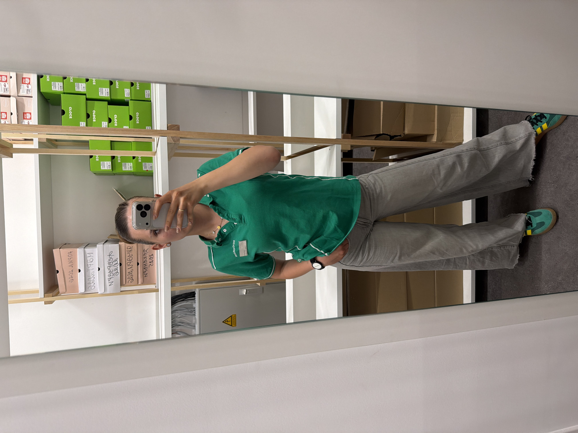 A person in a green collared shirt and gray jeans takes a photo of themself in a mirror. The mirror is in a room with shelves filled with boxes of shoes labeled "LOC2," "CLOC2," and "CLOCE." Other boxes are labeled with dates, including "26.05," "28.08," "JGKGHPON," "2606796M," and "HUDNY TOS." There is a DEICHMANN name tag on the individual's shirt.