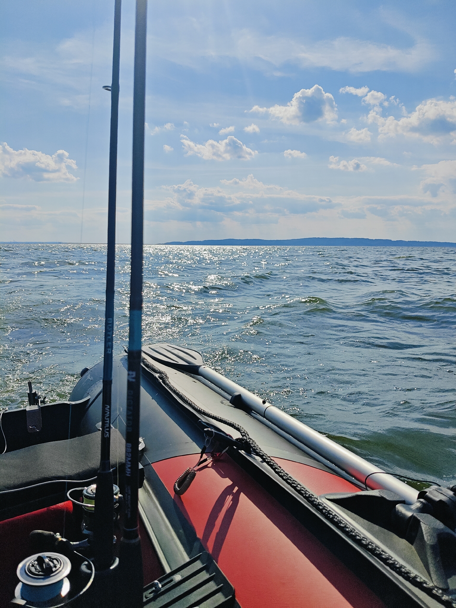 The image depicts a view from a boat on a body of water, likely the Kama River, in the vicinity of Chaykovsky, Russia. Two fishing rods, one with the brand "NAUTILUS" visible, are positioned upright in the foreground, resting on the interior of the boat. A fishing reel is also visible attached to one of the rods. An oar with a black grip and a metallic shaft is secured along the side of the boat. The boat itself appears to be inflatable, with red and black sections visible. The water is rippled, with sunlight glinting off the surface, suggesting a bright, clear day. In the distance, a tree-lined shore or island is visible on the horizon. The sky is blue with scattered white cumulus clouds.