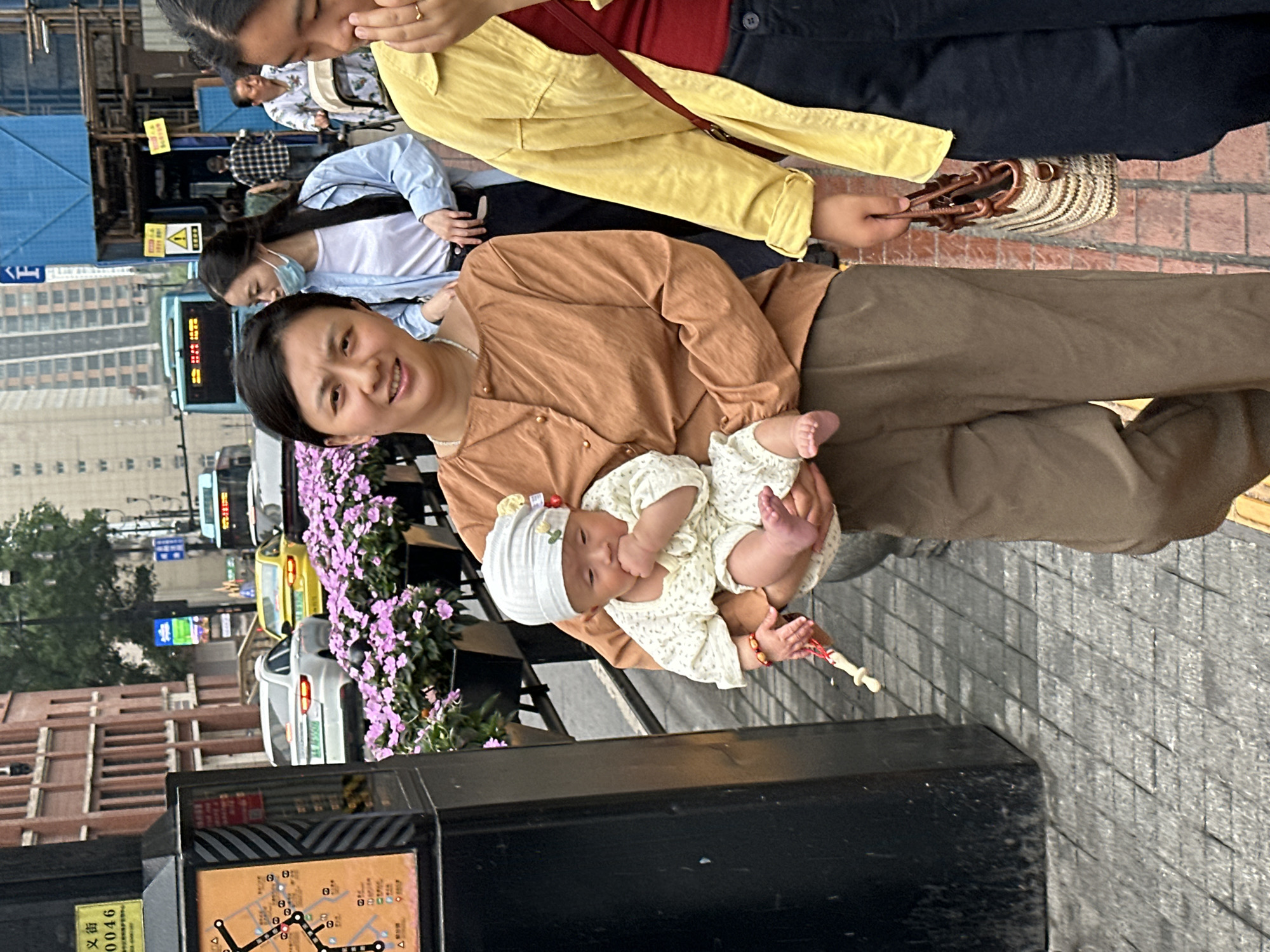A street-level, full-frame shot shows a woman holding a baby in Yuzhong District, China. The woman, wearing a brown shirt and pants, smiles at the camera while cradling a baby dressed in a white, floral-patterned outfit and matching hat. The baby is holding a white object with red and white string wrapped around it.
The woman is standing on a gray-paved sidewalk next to a black metal structure displaying a transit map. Behind her, a row of purple flowers grows along the sidewalk. In the background, there are several cars, a bus, and tall buildings, indicating an urban setting. Other people are walking nearby. A woman with a yellow jacket and straw handbag stands to the right. A woman in a light blue shirt stands behind the first woman.