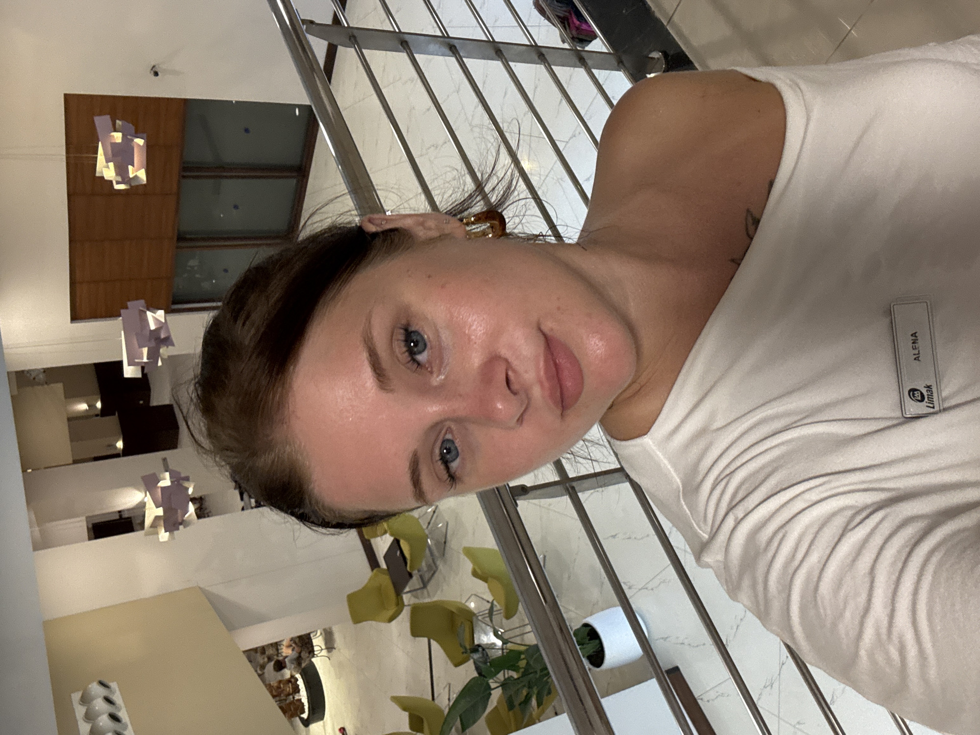 A woman with light skin, blue eyes, and brown hair is taking a selfie on a staircase in a hotel lobby. She is wearing a white t-shirt that is off one shoulder, revealing a tattoo on her shoulder. She has hoop earrings in her ears. A name tag is visible on her shirt with "ALENA" and "Limak" written on it. The hotel lobby has a modern design with white marble floors, yellow chairs, and hanging light fixtures. The image also shows a railing for the staircase and a large window in the background. The location is the Limak Lara De Luxe Hotel in Aksu, Turkey.