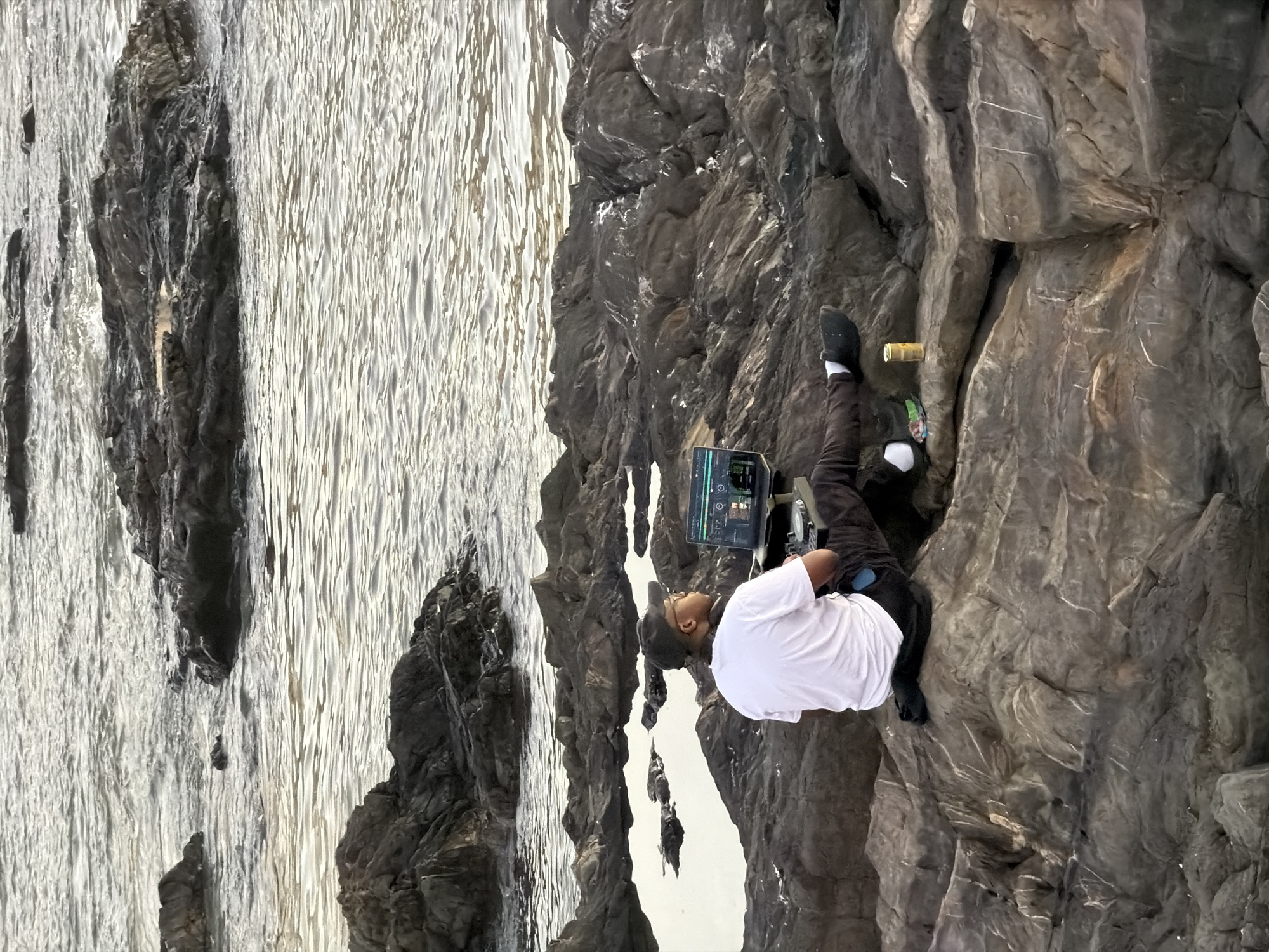 A man wearing a white T-shirt, black pants, and black shoes is suspended on a rocky cliff face. He is wearing a black cap and is looking at a screen attached to his waist. The background shows water and more rock formations. The scene appears to be outdoors in a natural environment. The location is on Chapman's Peak Drive, near Cape Town, South Africa.