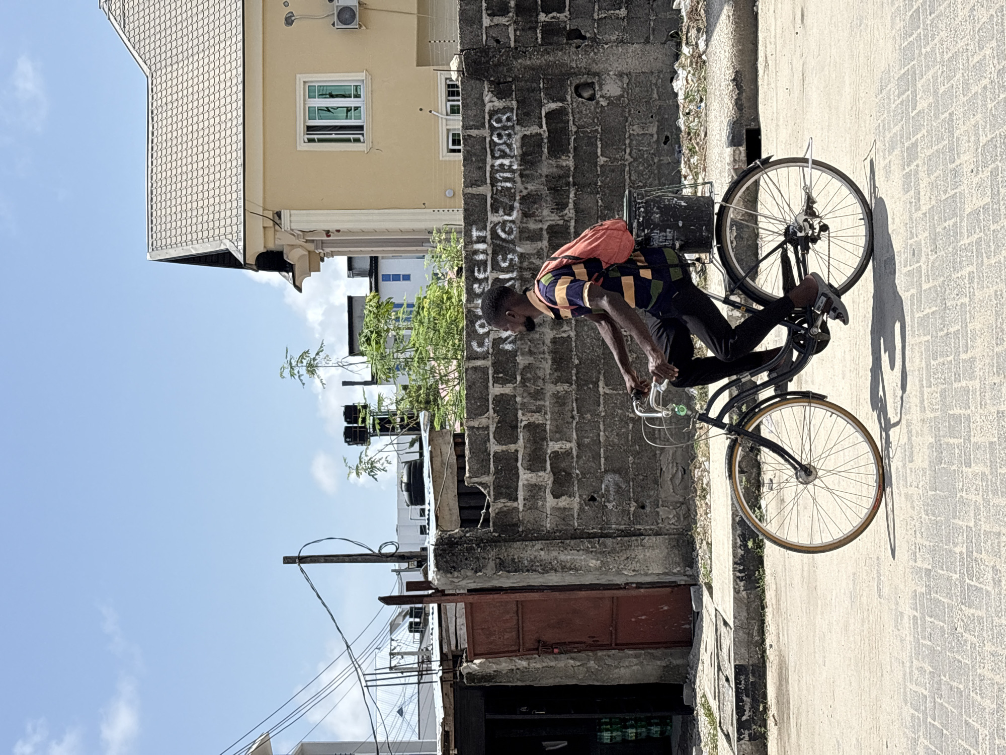 This image depicts a sunny street scene in Igboefon, Nigeria, featuring a man on a bicycle. The man, with dark skin, short dark hair, and a beard, is captured mid-action or paused on his bicycle. He is wearing a multi-colored, striped short-sleeved shirt (featuring blue, yellow, green, and black), dark trousers, and an orange backpack. His hands are on the handlebars of a dark-framed bicycle that appears to have a rear basket.
The man is on a light-colored, paved pathway or street. To his left, a weathered concrete block wall runs parallel to the path. On this wall, white paint clearly spells out "COASSENT N I.S / GEL 713288", along with other less legible markings. Beyond the wall, a two-story light yellow building with a distinctive textured or patterned roof is visible. It features a white-framed window with green reflections and an outdoor air conditioning unit. Below this building and behind the wall, parts of a smaller, darker structure with a reddish-brown metal door can be seen.
The bright daylight casts strong, clear shadows of the bicycle and rider onto the pavement, suggesting it is either mid-morning or afternoon. The sky is predominantly clear with scattered white clouds. Green foliage from trees or bushes is visible in the background, and a utility pole with wires extends into the frame from the lower left, indicating an urban or semi-urban setting. The scene conveys a sense of daily life and activity.