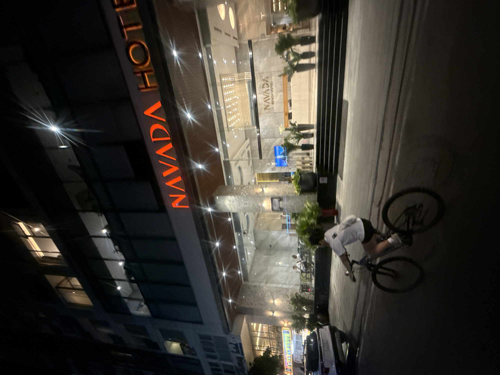 At night, a person rides a bicycle on the dark asphalt road in front of the Navada Beach Hotel in Bac Nha Trang Ward, Vietnam. The hotel's illuminated signage displays "NAVADA HOTEL" in bright red letters. The hotel features a modern facade with large glass windows and a well-lit entrance area. Several potted plants and decorative urns are placed on the steps leading up to the hotel's lobby, where a reception desk is visible. The streetlights and internal building lights cast reflections on the wet pavement. A white car is parked on the left side of the road. The sky is dark, indicating it is nighttime.