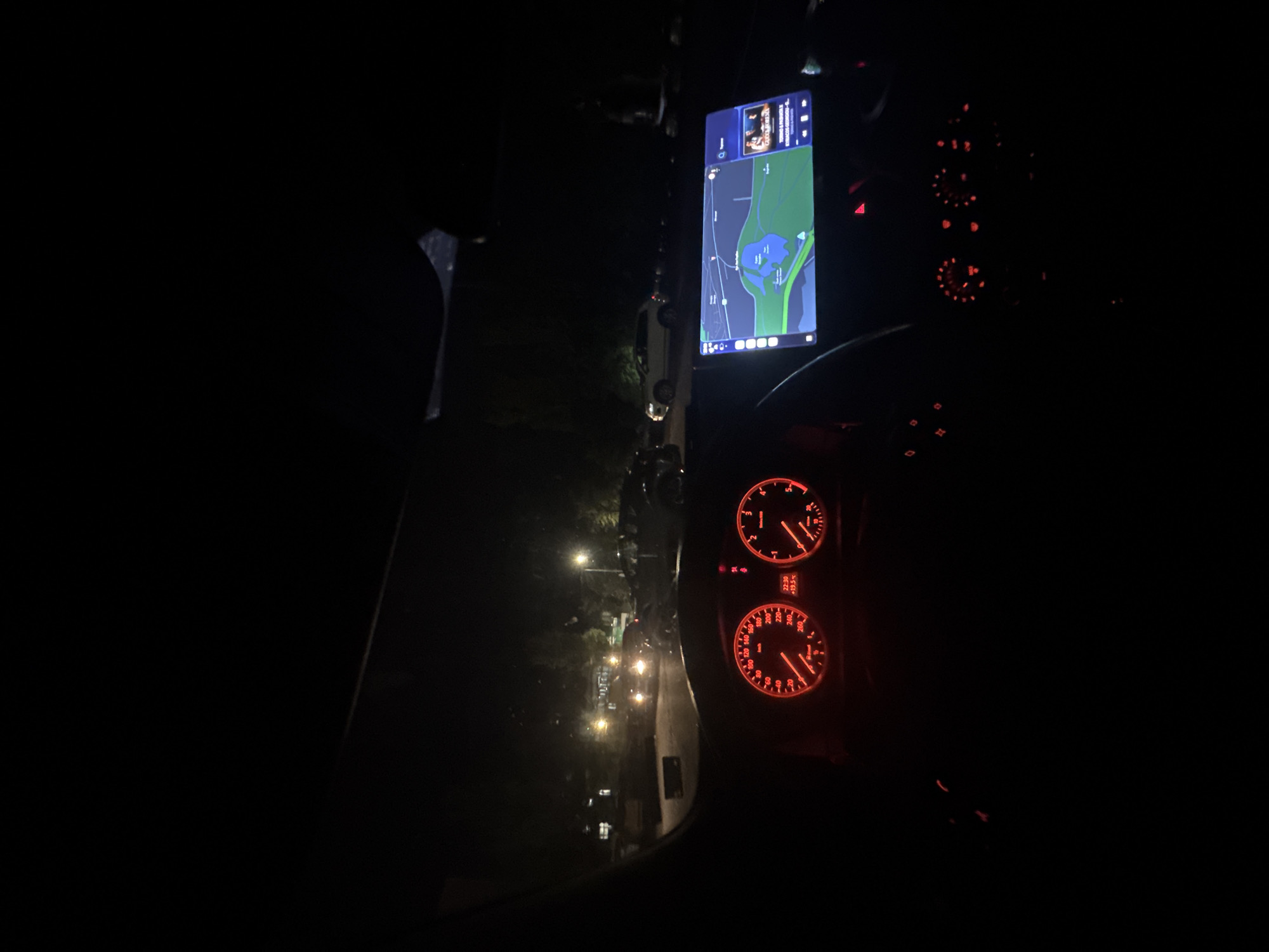 The image is taken from inside a vehicle at night, likely a car, as indicated by the dashboard instrumentation visible. A navigation system or phone screen is mounted on the dashboard displaying a map interface, with a route overlayed. Based on the map displayed on the screen, the location depicted appears to be in Stara Zagora, Bulgaria. The view through the windshield reveals a dimly lit street, potentially a residential area, with streetlights and other vehicles parked alongside the road. No people are clearly visible, and the focus is primarily on the navigation display and the surrounding environment viewed from within the vehicle. The dashboard indicates the time as 22:30, and the ambient temperature is +19.5°C. The overall lighting is dark, with the interior illuminated by the red glow of the dashboard lights and the screen.