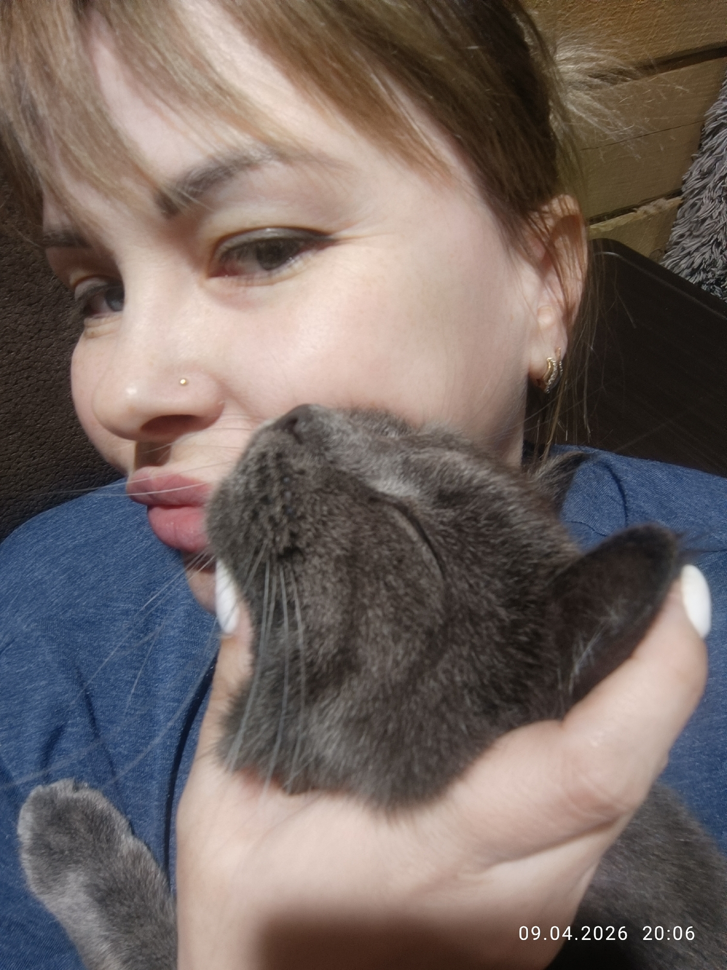A woman with light hair and a nose piercing is holding a gray cat. The cat's face is near the woman's face. The woman is wearing a blue shirt. The photo was taken on April 9, 2026, at 20:06. The location is likely Chaikovsky, Russia.