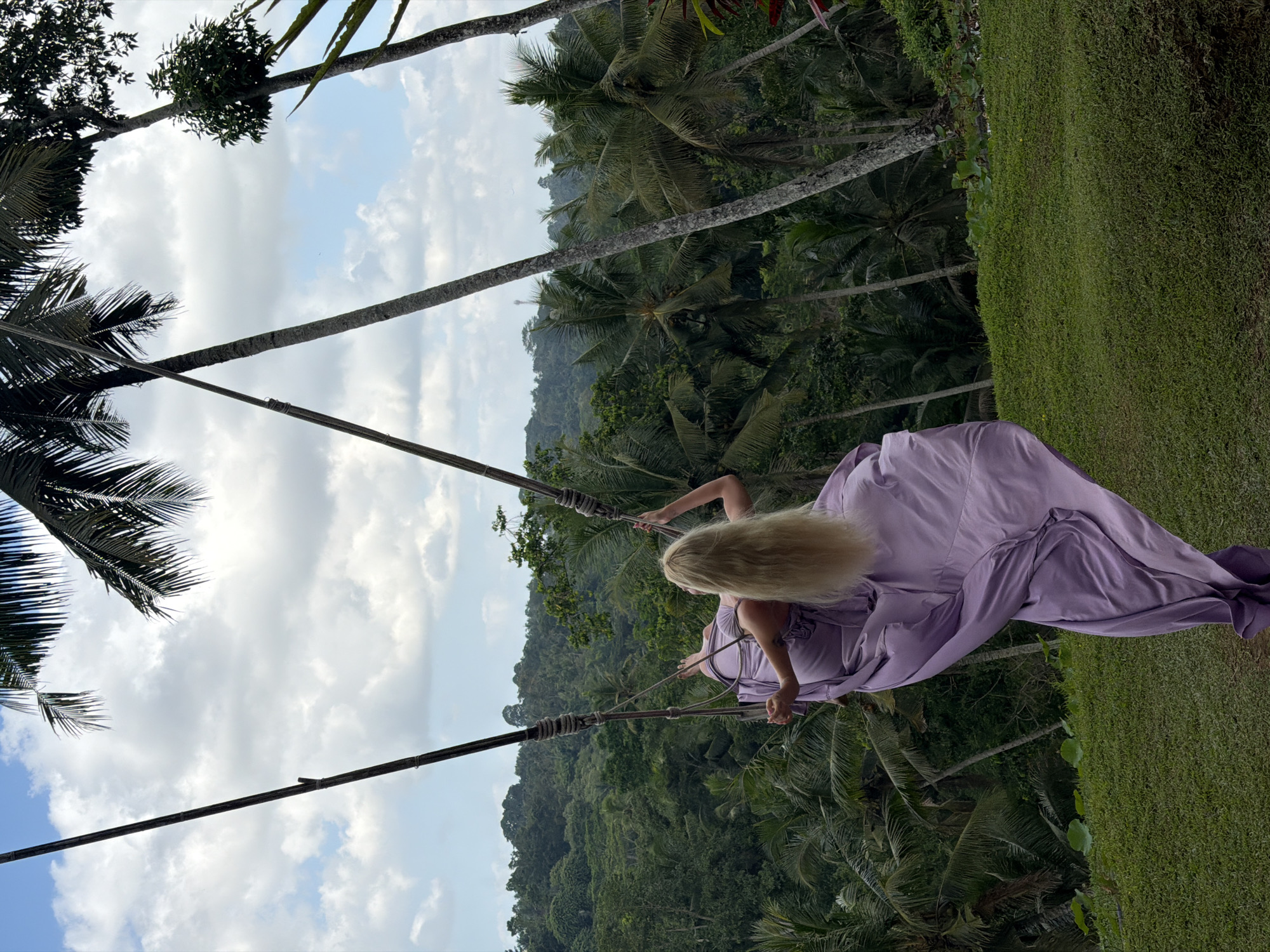 A blonde woman with long hair is swinging on a rope swing. She is wearing a long, flowing lavender dress that billows around her. The swing is suspended from thick ropes attached to two wooden poles. The background is a lush, green jungle with numerous palm trees and a backdrop of rolling hills under a cloudy sky. The woman is facing away from the viewer, looking out at the jungle. The scene appears to be an outdoor tourist attraction.