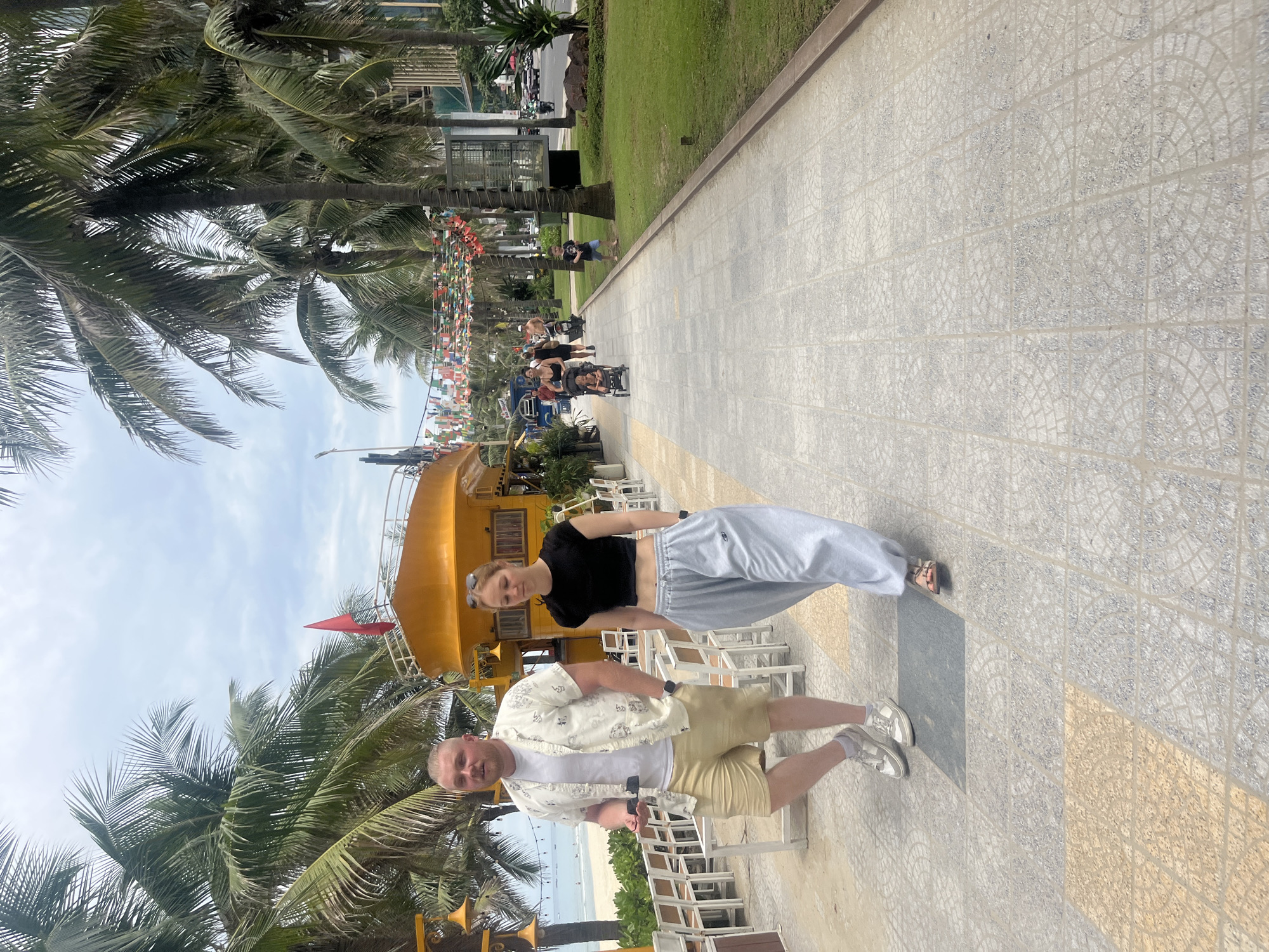 In Đà Nẵng, Vietnam, a man and a woman walk along a paved pedestrian pathway lined with palm trees and outdoor seating. The man, in a light-colored patterned short-sleeved shirt, white t-shirt, and beige shorts, walks to the left. The woman, in a black crop top and baggy grey pants, walks to his right, slightly ahead. Behind them, a yellow vehicle resembling a boat on wheels is parked. Further down the pathway, other people are visible, some with strollers, and a child stands on the grass to the right. The environment suggests a beachfront area with a bright, cloudy sky.