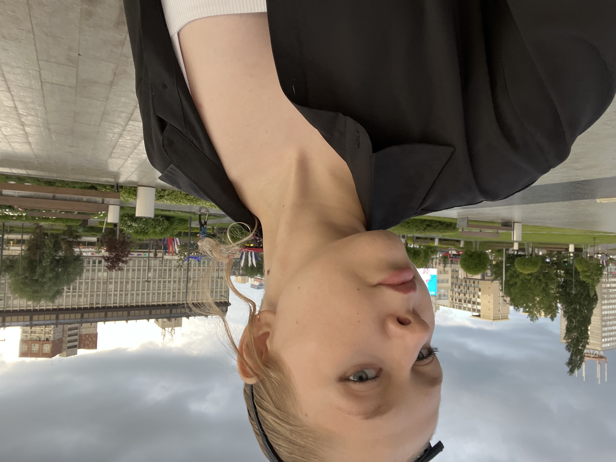 A young Caucasian woman with blonde hair pulled back by a black headband and wearing a black open button-up shirt over a white tank top is looking towards the camera. Her face is turned slightly to the left. The background shows an urban environment with modern buildings and trees under a cloudy sky. A paved walkway and park benches are visible in the foreground. The image appears to be taken in the city of Perm, Russia, possibly near a central square or park area.