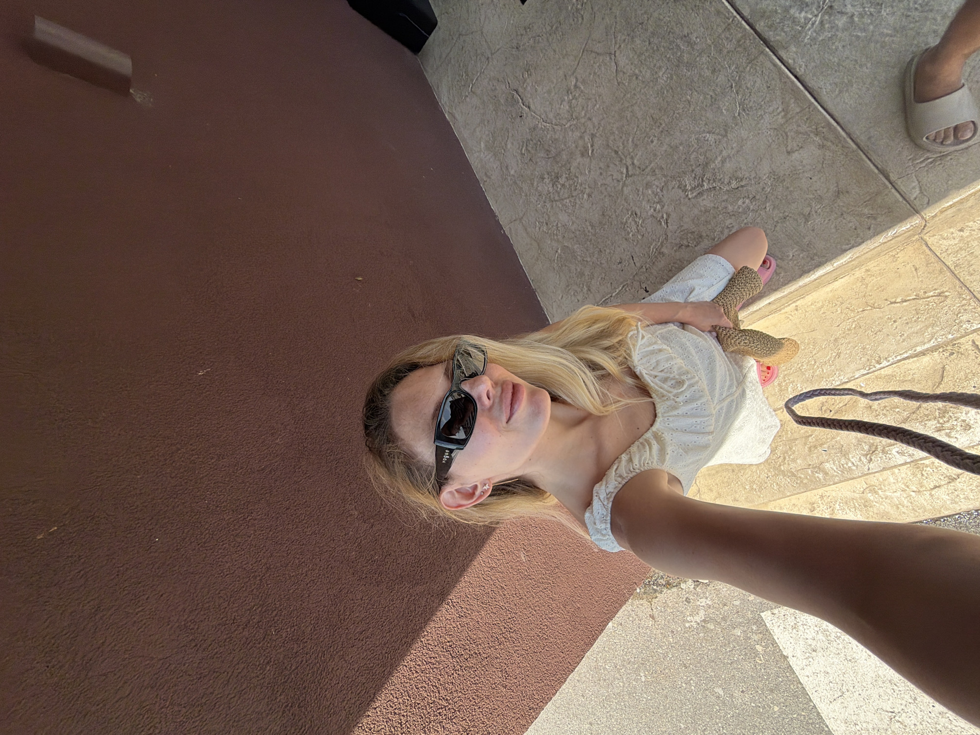 The image shows a young woman with blonde hair and sunglasses kneeling on a textured stone surface. She is wearing a white off-the-shoulder dress and holding a straw bag. Her right leg is extended, and her left knee is bent. The background consists of a brown textured wall and a shadow cast by an unseen structure. The scene appears to be outdoors, possibly in a public space given the stone flooring and wall. The lighting suggests it is daytime, with sunlight casting distinct shadows. There are no clear indicators of the specific location within Bigor, Montenegro.