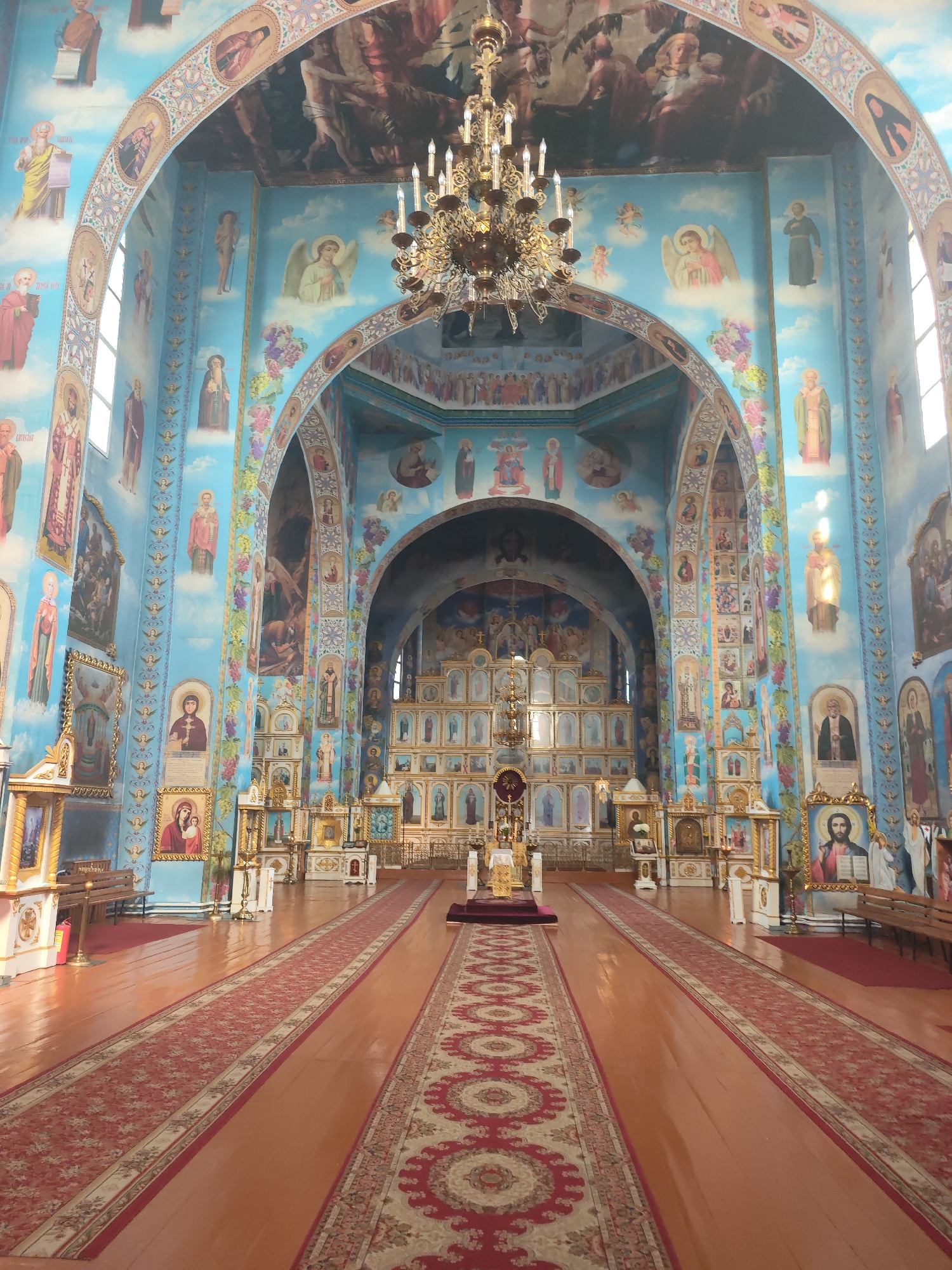 The interior of the Church of Saints Peter and Paul in Slonim, Belarus, is depicted in this image. The church is ornately decorated with religious iconography covering the walls and ceilings. A grand chandelier hangs from the vaulted ceiling, and rows of pews are visible on either side of a long, red carpet leading to the altar. The altar itself is adorned with icons and religious symbols. The overall atmosphere is one of solemnity and religious devotion.