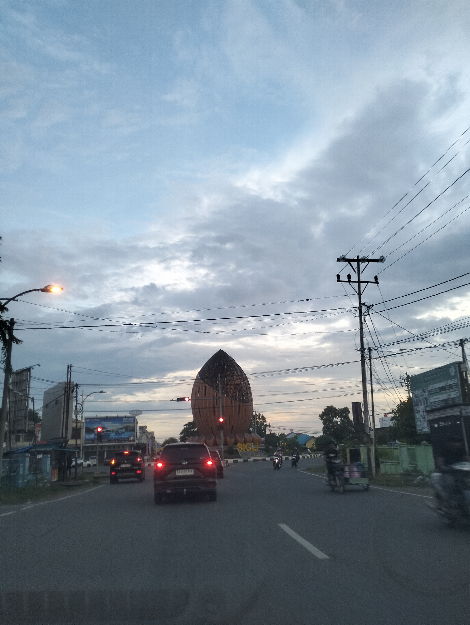 The image captures a street scene in Sigli, Indonesia, during twilight. The sky is overcast with diffused light, indicating either early morning or late evening. A prominent, tall, brown, dome-shaped structure, possibly a landmark or monument, stands on the right side of the road, adorned with the word "SIGLI" in large letters at its base. A traffic light with a red signal is visible near the structure.
The road in the foreground is asphalt, with a white line marking the lane. Several vehicles are present: a dark SUV in the center lane, its brake lights illuminated, and other cars and motorcycles further down the road. Some vehicles appear to be moving, while others are stopped at the traffic light.
On the left side of the road, there are buildings, streetlights, and utility poles with numerous wires crisscrossing the sky. A bright streetlight illuminates the immediate area on the left. On the right side of the road, there are more buildings, a green fence, and trees.
Several individuals are visible, mostly on motorcycles. One person is operating a motorcycle in the right lane, while others are further in the distance, moving along the road. The overall atmosphere is that of a typical urban street during a transitionary period of daylight.