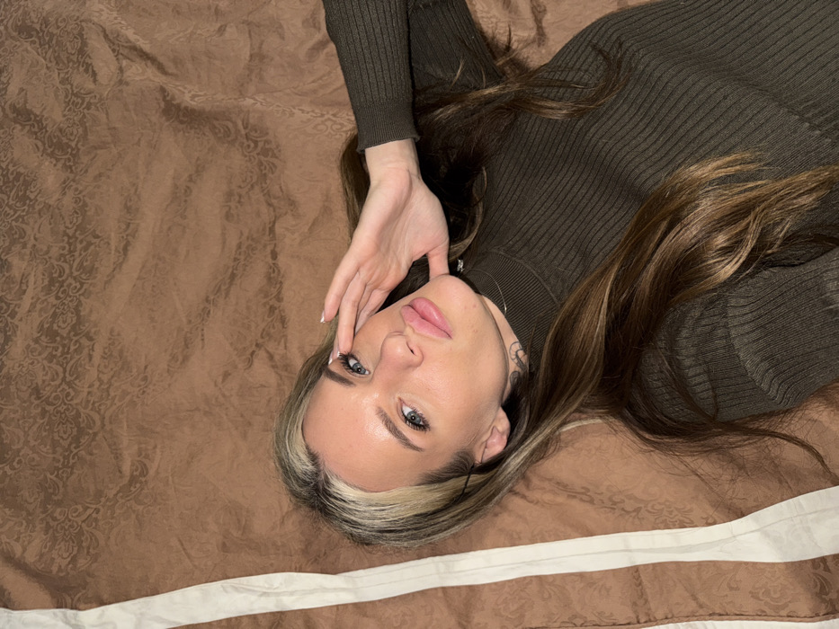 A fair-skinned woman with striking blue eyes lies on her back on a patterned brown comforter with a white-trimmed edge. She wears a fitted, dark green, long-sleeved top. Her long brown and blonde hair is spread around her. She has a tattoo on her neck. Her right hand is raised near her face, palm facing inward. Her expression is neutral, with pink lips slightly parted.