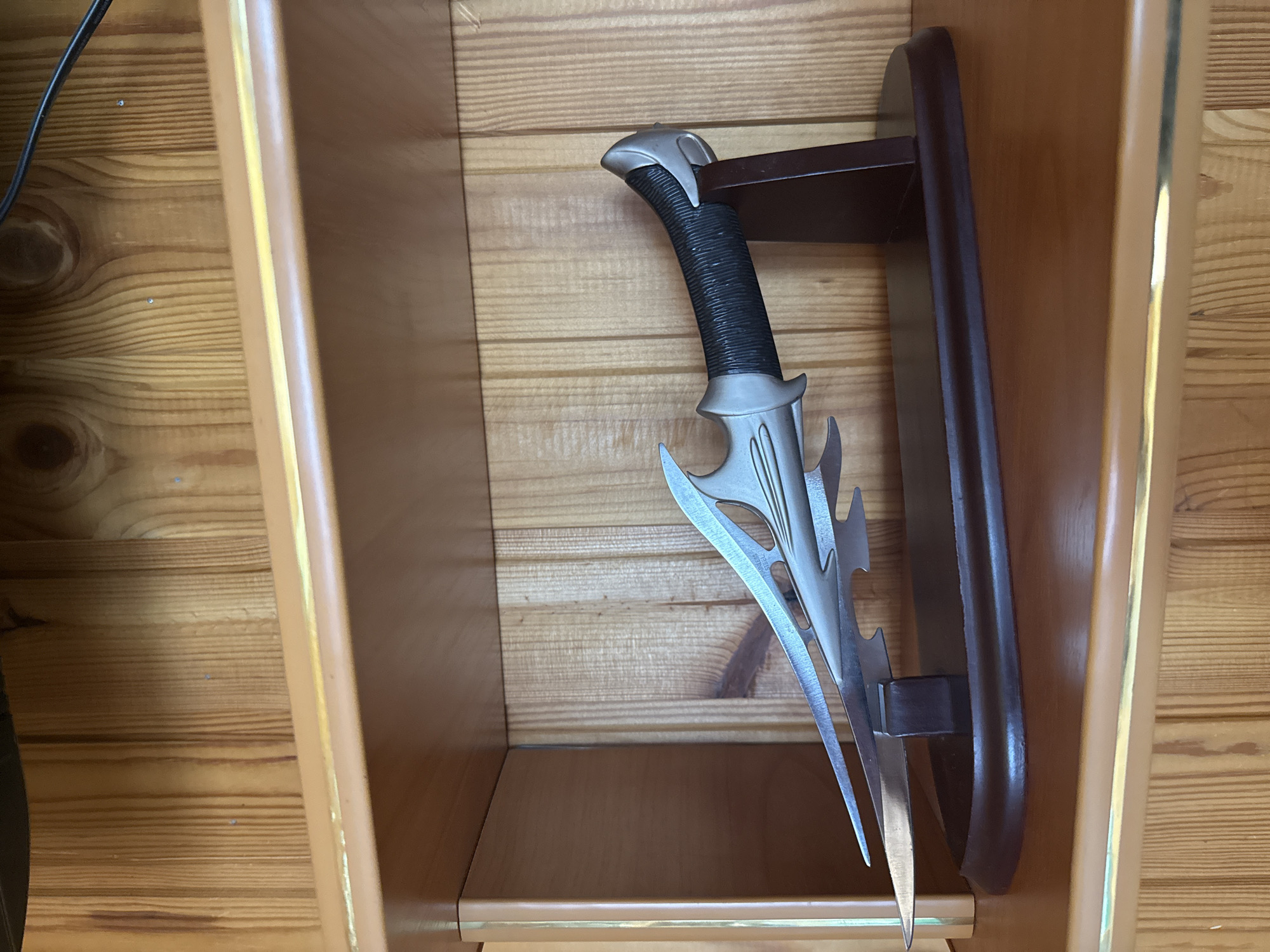 A decorative dagger with a silver blade and a black, textured handle rests on a dark wooden display stand, which is positioned on a wooden shelf. The background consists of a light brown wooden paneled wall. The dagger's blade is elongated and features sharp, pointed protrusions along its length, with several cut-out sections. The handle is wrapped tightly with dark material, and the pommel is a metallic silver cap. The display stand is oval-shaped with a dark wood finish and has a vertical piece supporting the dagger's handle. The shelf is made of polished wood and has a light-colored trim along its edge.