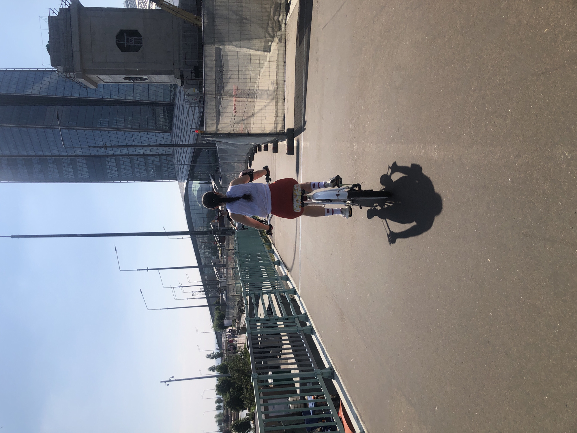 A woman is riding a bicycle on a paved pathway during the daytime. She is wearing a white sleeveless top, red shorts, white socks with red stripes, and black sneakers. She has a black ponytail and is wearing black wristbands. The bicycle is white with black tires. Her shadow is cast on the pathway behind her. In the background, there are tall modern buildings, a bridge with a light blue railing, streetlights, and some construction fencing. The sky is clear and blue. The scene appears to be in an urban environment.