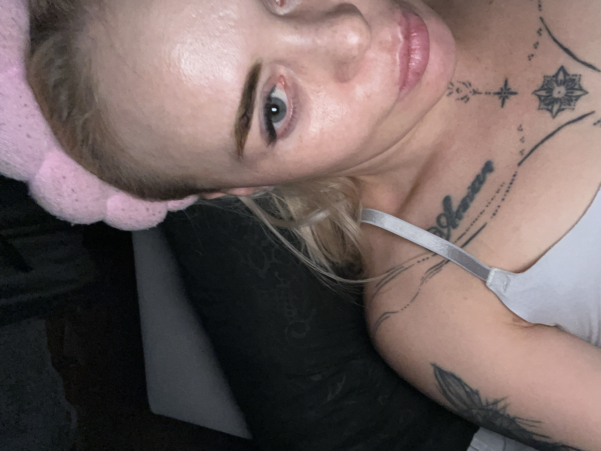 The image shows a close-up, selfie-style shot of a fair-skinned woman with blonde hair. She is wearing a pink fuzzy headband and a white bra. She has several tattoos, including one on her upper arm and chest. Her eyes are open and looking at the camera, and her lips are slightly parted. The background is dark and appears to be indoors, with a hint of a dark-colored pillow or cushion behind her. The lighting suggests it may be nighttime or a dimly lit room. No city or location information is discernible from the image itself.