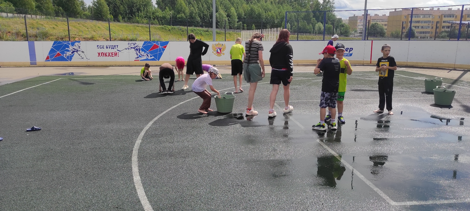 In Noviy, Russia, on a fenced outdoor sports court with a mural depicting hockey players and Russian text reading "Всё будет ХОККЕЙ!" (Everything will be HOCKEY!), several children are engaged in water-based activities under a partly cloudy sky. The court surface is wet, with puddles reflecting the surroundings. A group of children, varying in age, are scattered across the court. One child in the foreground, wearing a white t-shirt and brown pants, is bent over a green bucket, seemingly interacting with the water inside. Other children are standing, crouching, or walking around the court. One boy in a black t-shirt with a yellow graphic and black pants stands near two green buckets. Another group of boys in colorful shorts and t-shirts are clustered together, one wearing a red cap. The background shows a grassy slope with trees, a fence, and residential buildings in the distance. The scene suggests a recreational or playful event involving water on a warm day.