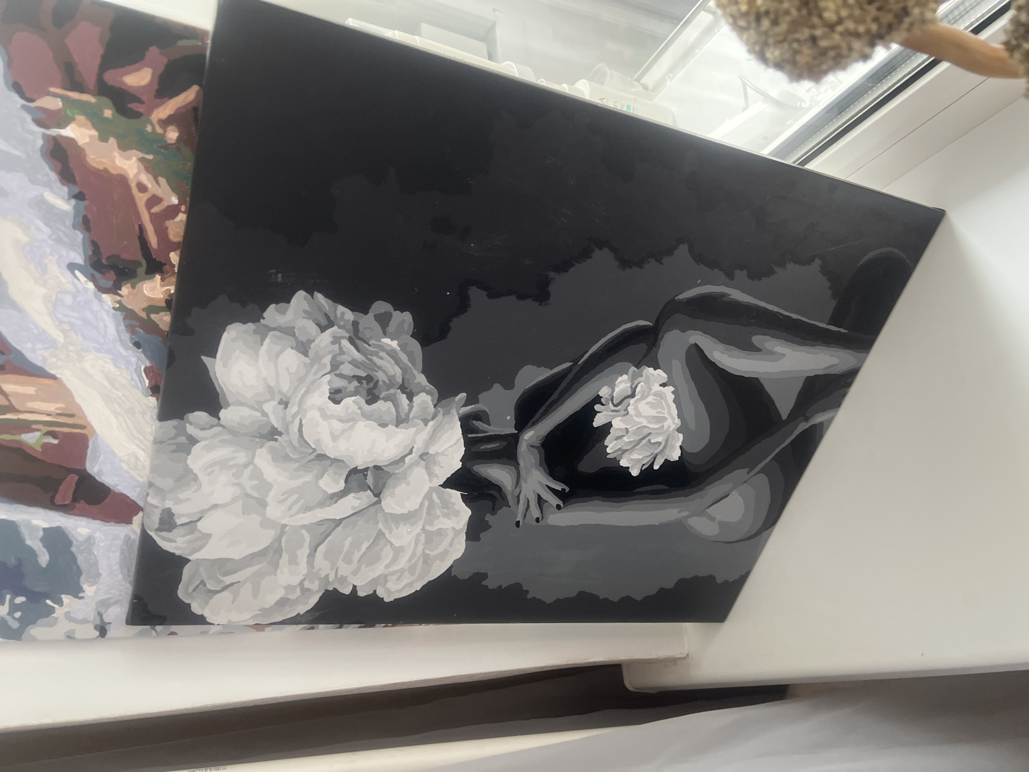 A greyscale painting depicts a woman with a large, light-colored flower for a head, sitting with her knees drawn up. She is holding a smaller, similar flower in her hands, which are resting on her lap. The background is dark grey. The painting is placed on a white windowsill, next to a sheer white curtain on the left and a window with a view of what appears to be a cityscape or buildings on the right. Another painting with colorful abstract shapes is visible behind and above the greyscale painting. A fuzzy brown object with a light-colored ear, possibly a stuffed toy, is positioned on the right side of the windowsill, partially obscuring the view outside the window.