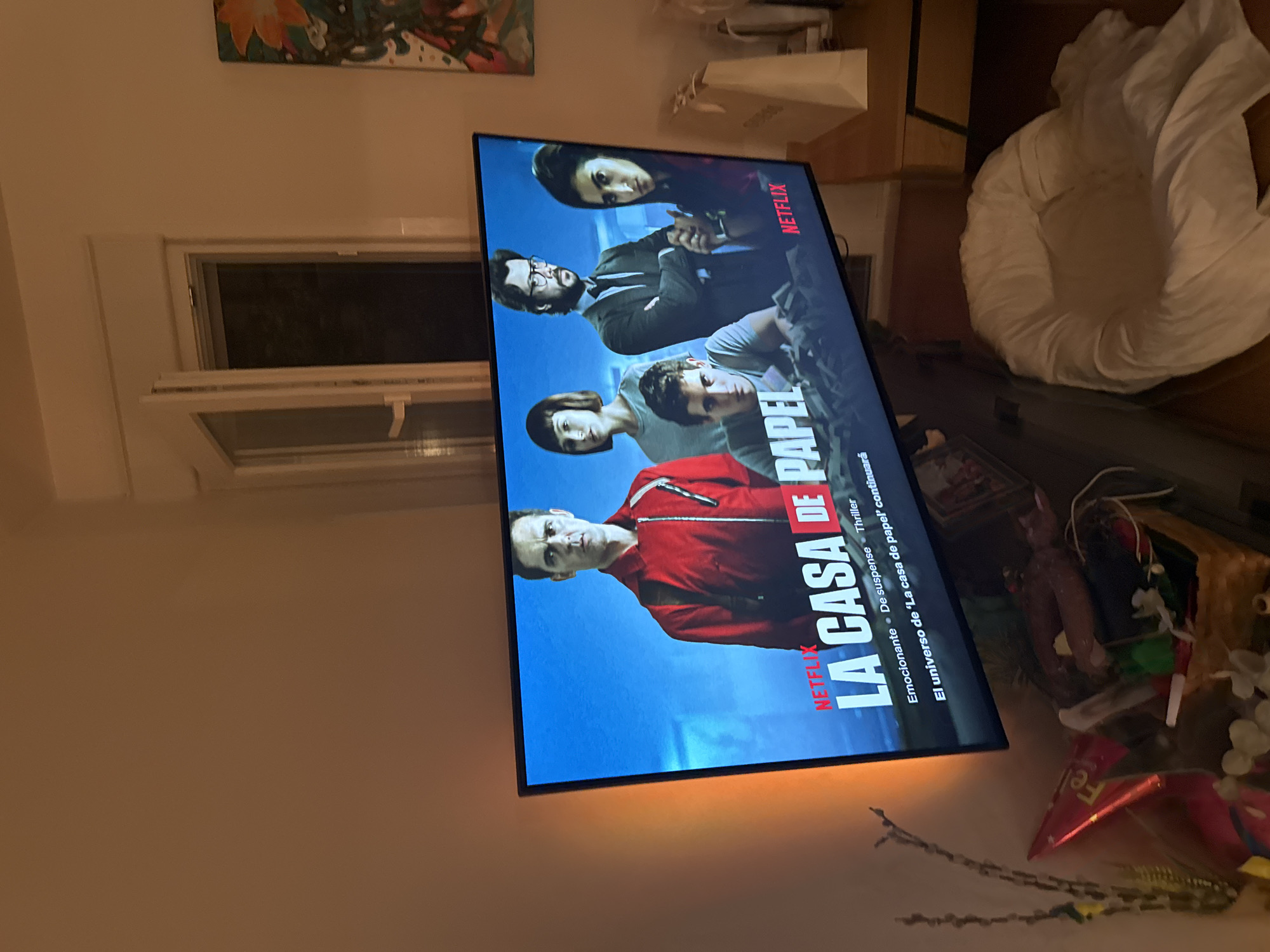 A television screen displays a promotional image for the Netflix series "La Casa de Papel" (Money Heist). The screen shows five characters: a man in a red jumpsuit, a woman with a short bob haircut, a man with glasses and a suit, a young man in a gray t-shirt, and a woman with dark hair. The title of the show is prominently displayed in large white lettering with red accents. Below the title, text reads "Emocionante De suspense Thriller" and "El universo de 'La casa de papel' continuará." The TV is angled slightly upwards and to the left. The background of the image is an indoor domestic setting. To the left of the TV, a wall is illuminated by an orange light source, casting a warm glow. To the right of the TV, a wooden table holds a white paper bag. In the foreground, near the bottom of the image, a woven basket is filled with various items, including what appear to be party decorations like a cone hat and possibly a doll. A large, white, plush beanbag chair is situated to the right of the basket. The overall scene suggests a living room environment, likely in a residential apartment, and the provided context of Murcia, Spain, places the scene geographically.