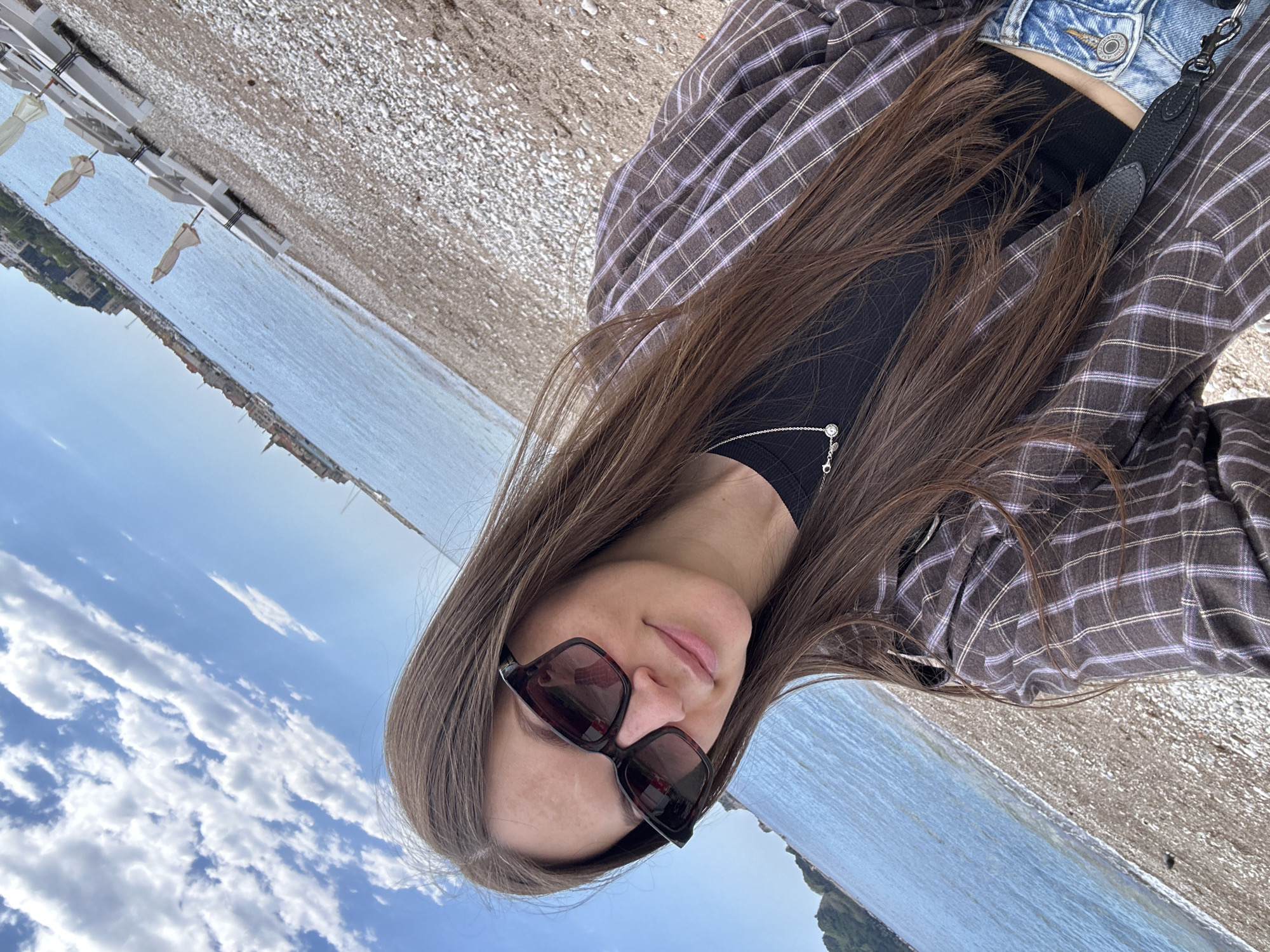 A woman with long brown hair, wearing sunglasses and a plaid shirt over a black top, poses for a selfie-style photograph on a beach. The photograph is rotated counter-clockwise. Behind her, the Adriatic Sea stretches towards the cityscape of Budva, Montenegro, visible in the distance. White beach umbrellas and lounge chairs line the beach, partially obscured by the camera angle. The sky above is blue with scattered white clouds. The woman appears to be standing on a pebble beach.