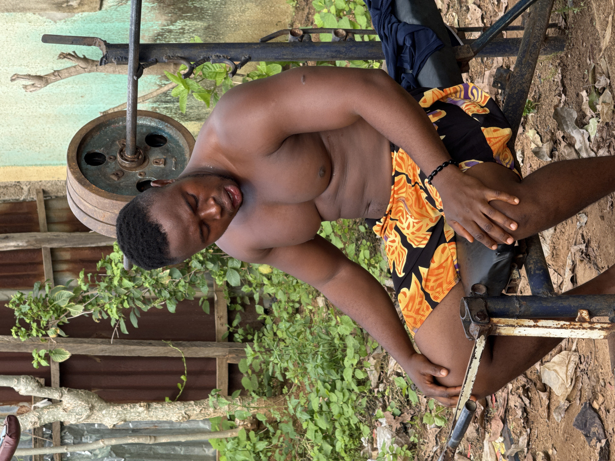 A shirtless Black male is pictured outdoors in a seated position, resting on what appears to be a weightlifting bench. He is wearing a pair of black shorts with an orange and yellow floral pattern. His arms are visible, one resting on his leg, the other near his chest. The scene is set in a somewhat rustic, outdoor environment with greenery and wooden structures in the background. A weight plate from a barbell is visible to the left of the frame, suggesting the activity is related to weightlifting or exercise. The ground is covered with dirt, fallen leaves, and some debris. The lighting suggests it is daytime. The man's facial expression is one of exertion or discomfort. The overall impression is of an informal, possibly improvised, outdoor gym setting.