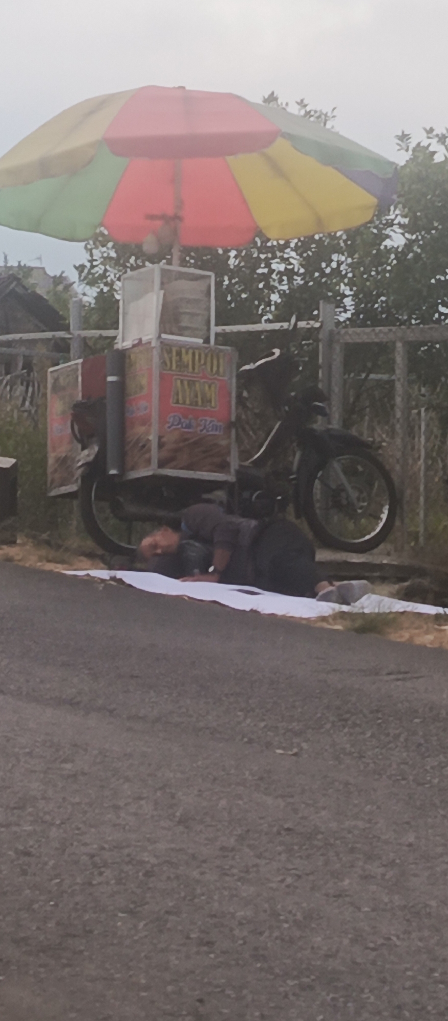A man is lying on his side on a white cloth on the side of a road in Sumbersekar, Indonesia. He is wearing a dark hoodie and dark pants, with a watch on his left wrist. Next to him is a motorcycle with a food cart attached. The food cart has an umbrella attached above it, which is red, yellow, and green. The cart has text that reads "SEMPOL AYAM" and "Pots Kim". The environment appears to be outdoors with a fence and trees in the background. The sky is overcast.