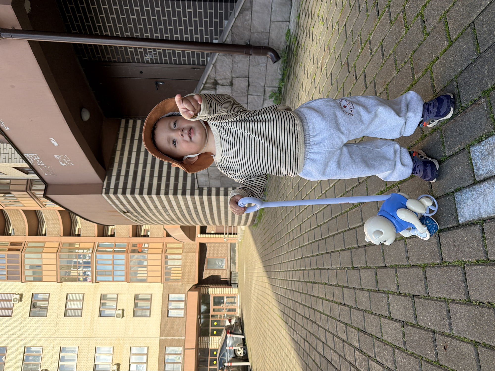 A young child, wearing a bucket hat, a striped long-sleeved shirt, and gray sweatpants, stands on a paved walkway in front of a modern apartment building. The child is holding onto the handle of a toy dog that rolls on wheels. The child is pointing to the right with their left hand. The apartment building is made of yellow brick and has multiple windows and balconies. A dark brown downspout runs along the side of the building. The walkway is made of interlocking gray pavers with grass growing in between. A car is parked to the left of the frame. The setting appears to be a residential area in the city of Akulovo (Gp Kubinka), Russia, based on the provided context.